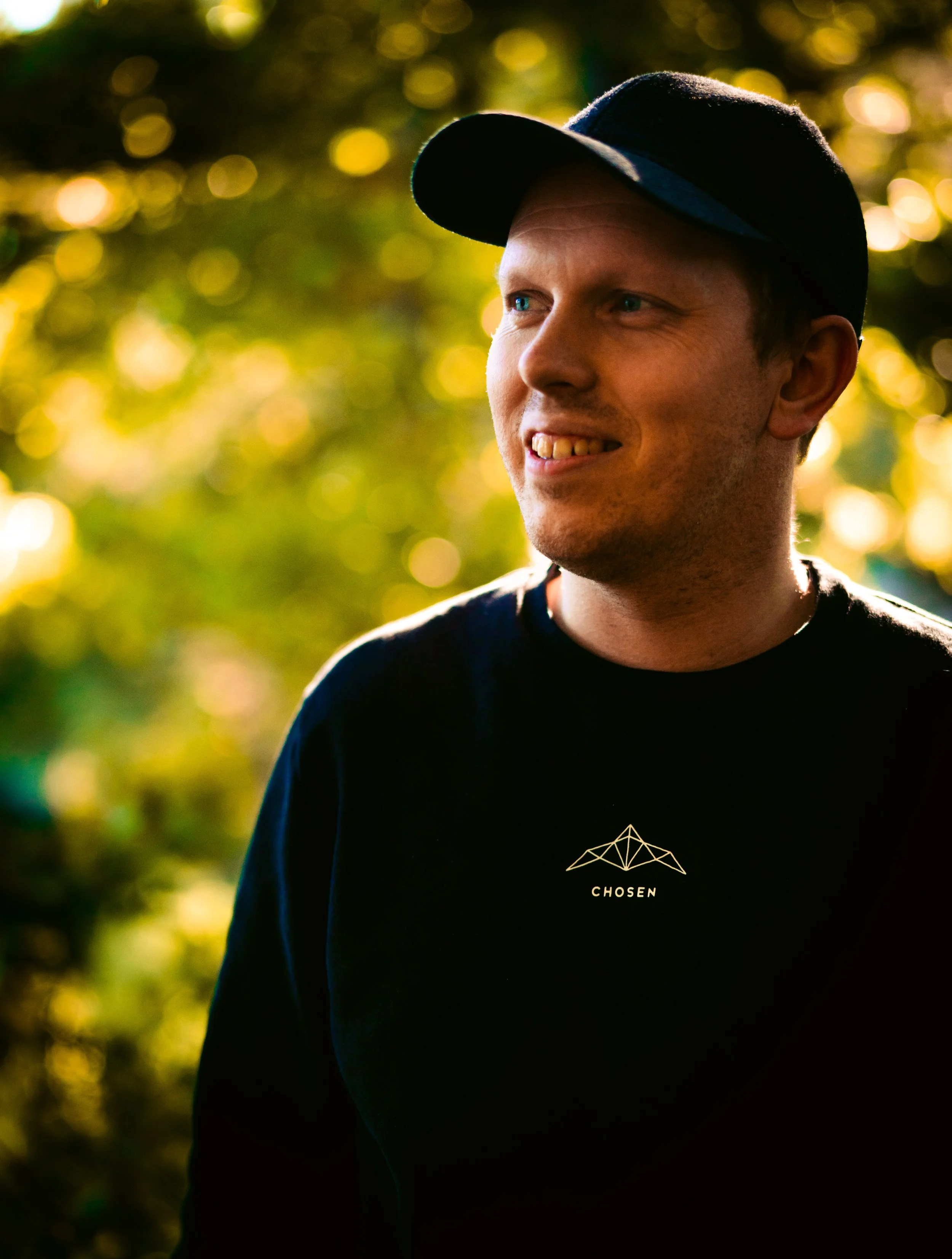 En mann med lue og svart T-skjorte som smiler, med sollys og grønne blader i bakgrunnen.