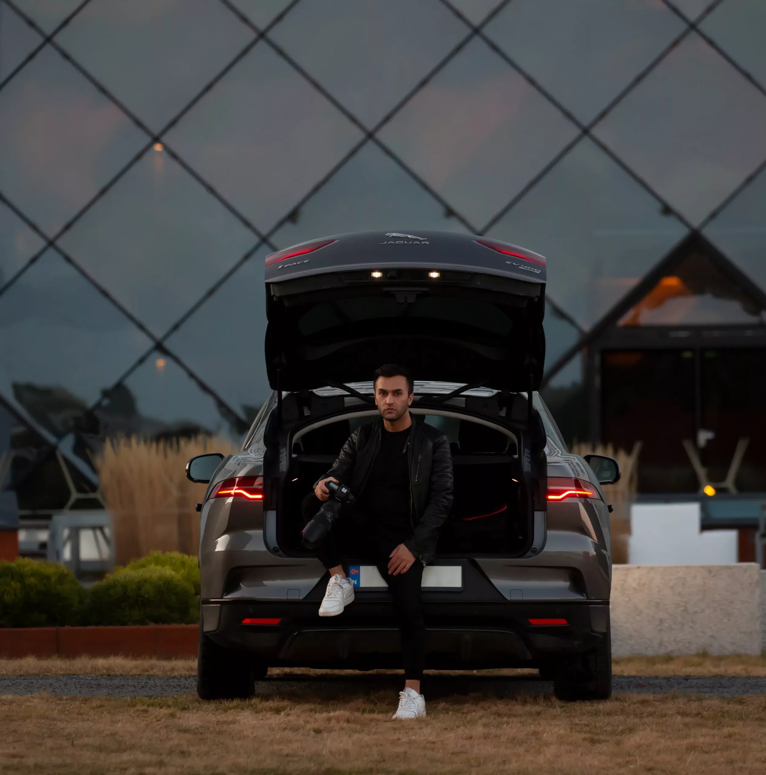 En mann i svart jakke og hvite sneakers sitter på fotbrønnen av en grå Jaguar elbil med åpen bakluke, mens han holder et kamera. Bakgrunnen viser en skyskraper med glassfasade og noe vegetasjon.
