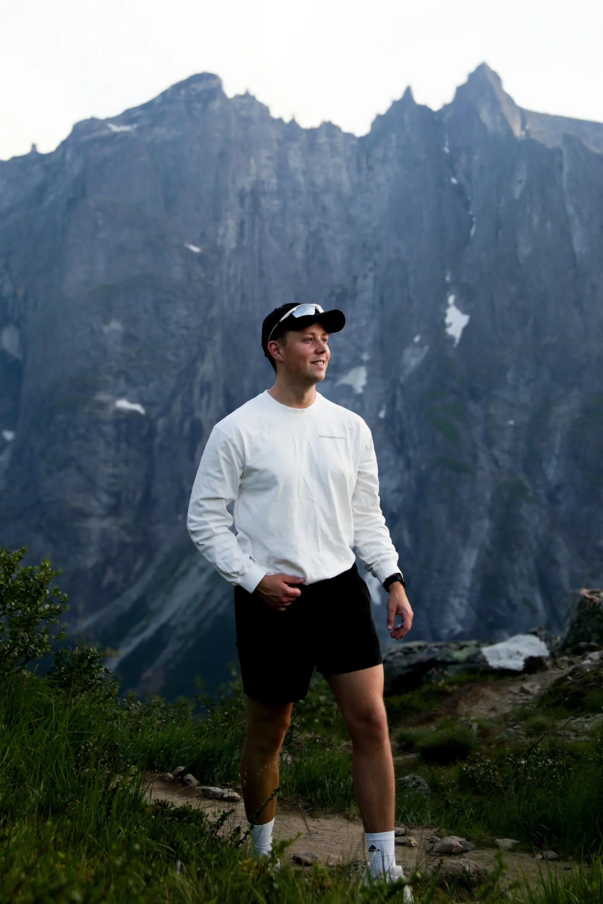 Ung mann på tur i fjellet, iført hvit jakke, sorte shorts, caps og solbriller, med fjell i bakgrunnen.