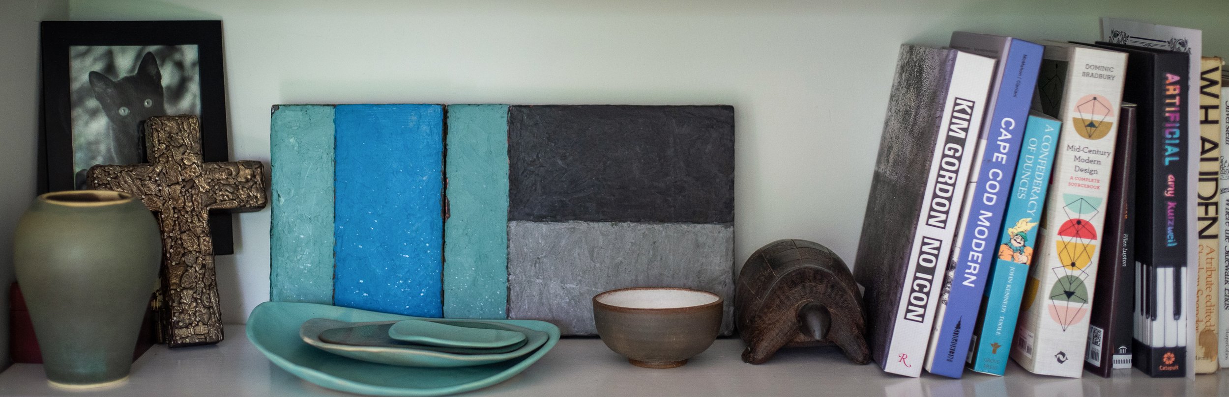 Decorative shelf with books, artwork, and pottery items including a framed black-and-white cat portrait, a gold cross, a green vase, and an assortment of colorful books.