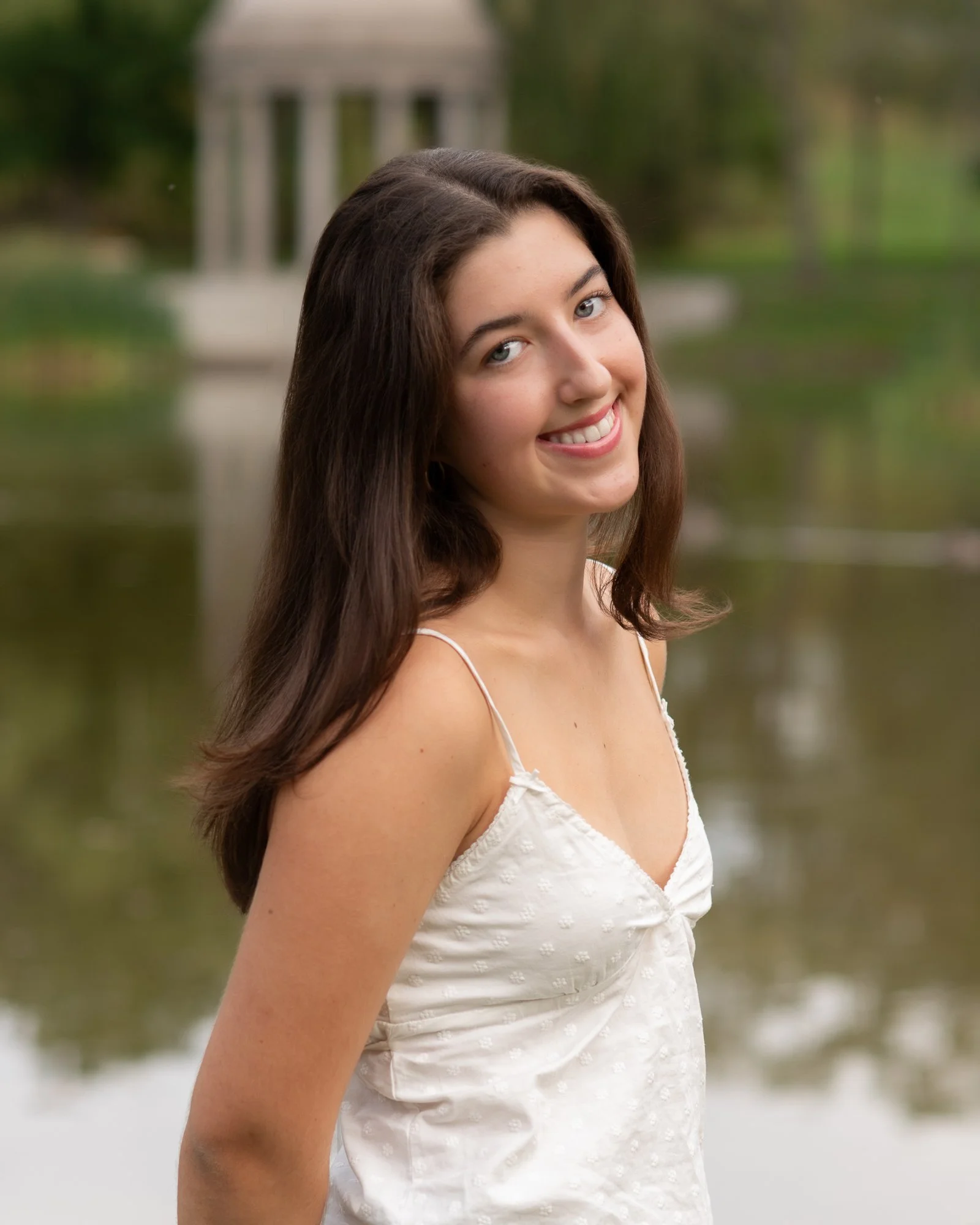 A young woman with long brown hair smiling outdoors near a body of water and a small gazebo in the background.