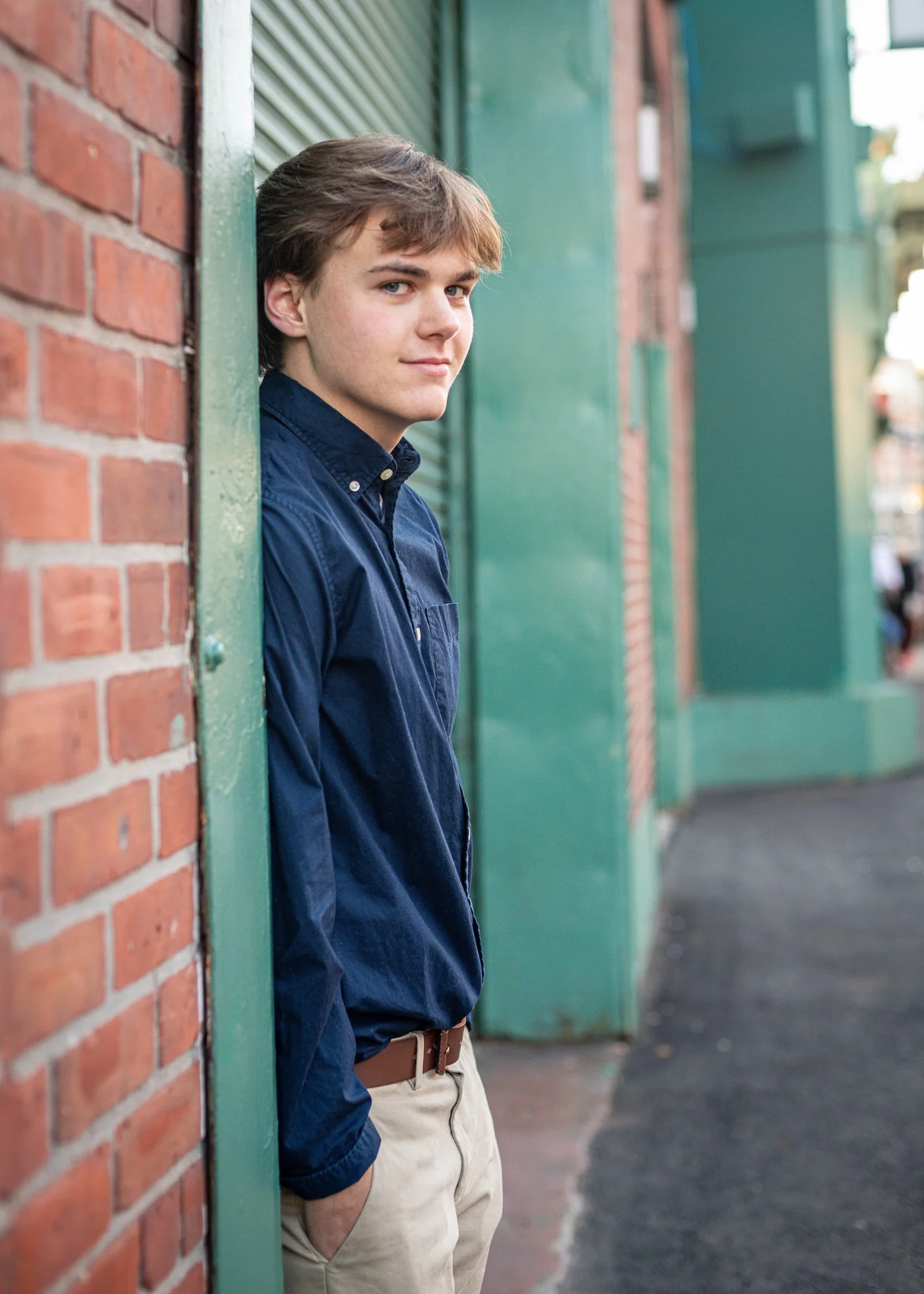 A young man with brown hair and blue eyes wearing a navy blue shirt and beige pants, leaning against a green wall on a city sidewalk.