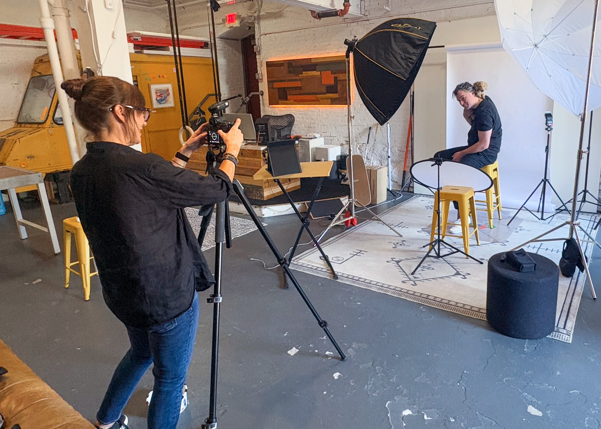 A woman getting filmed in a studio with a white backdrop, lighting equipment, and a person sitting on a yellow stool.