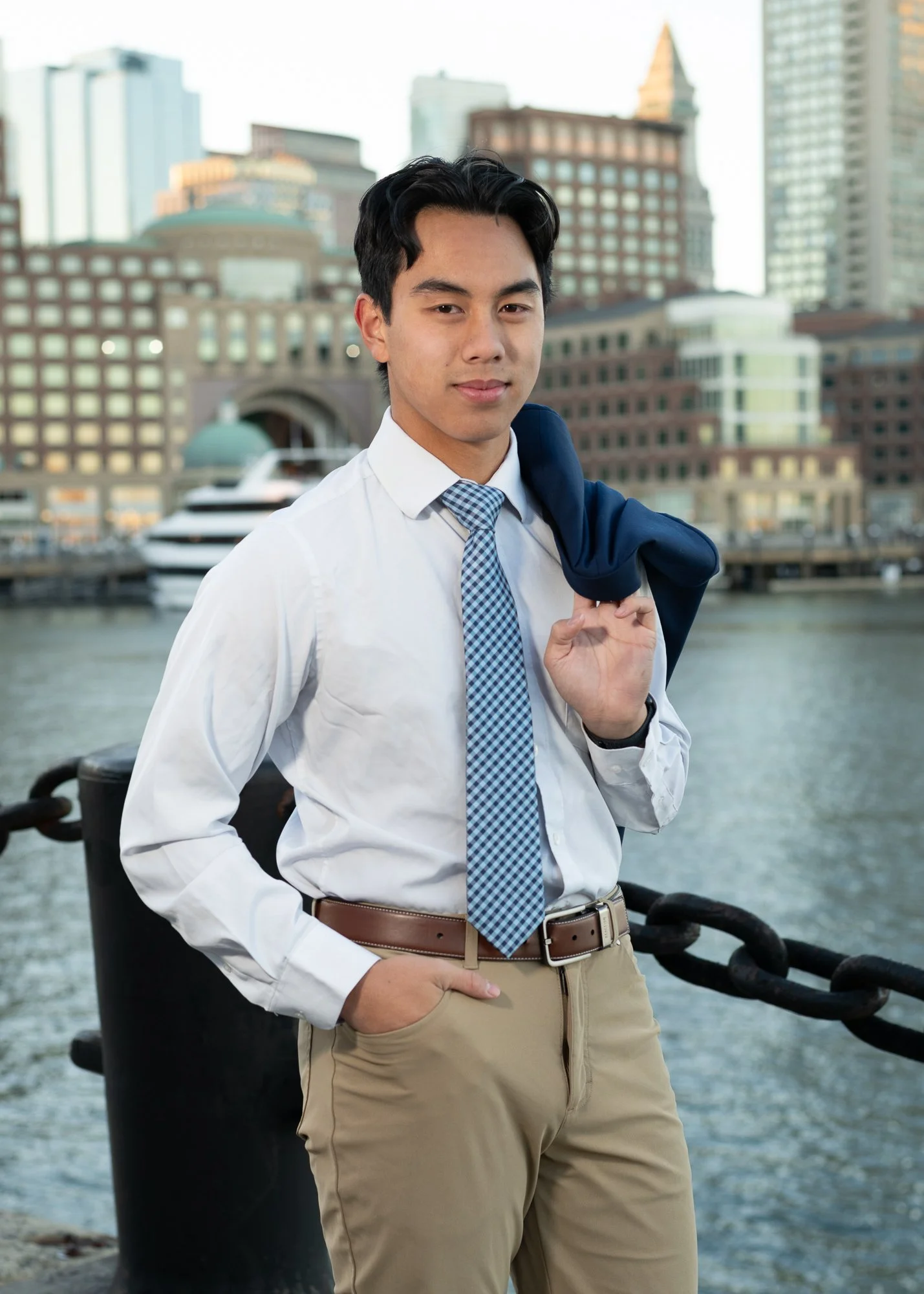 A young man in business casual attire standing outdoors near water with city skyscrapers in the background.