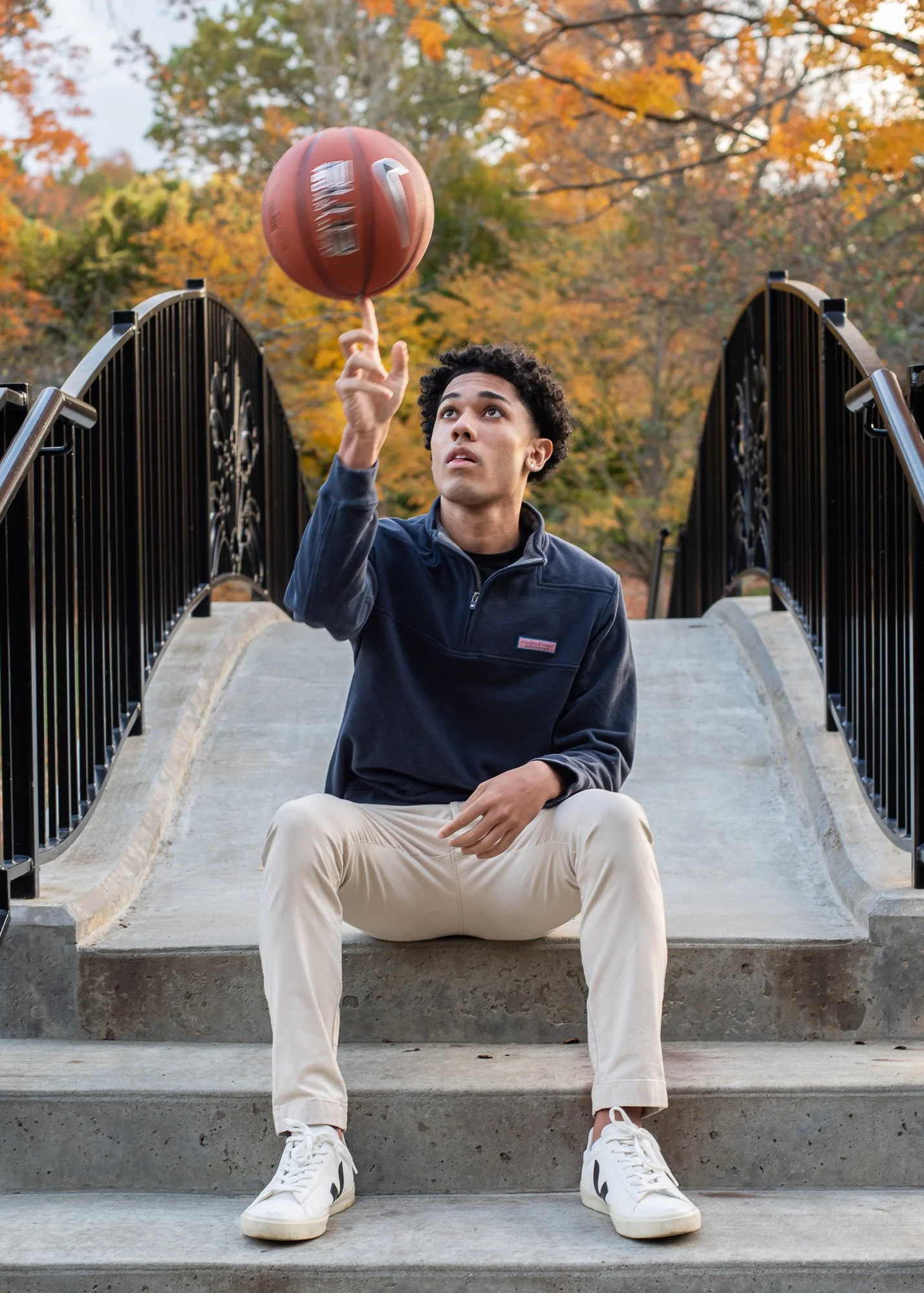A young man sitting on concrete stairs with steps and black metal railings, spinning a basketball on his finger outdoors during autumn, with colorful fall trees in the background.