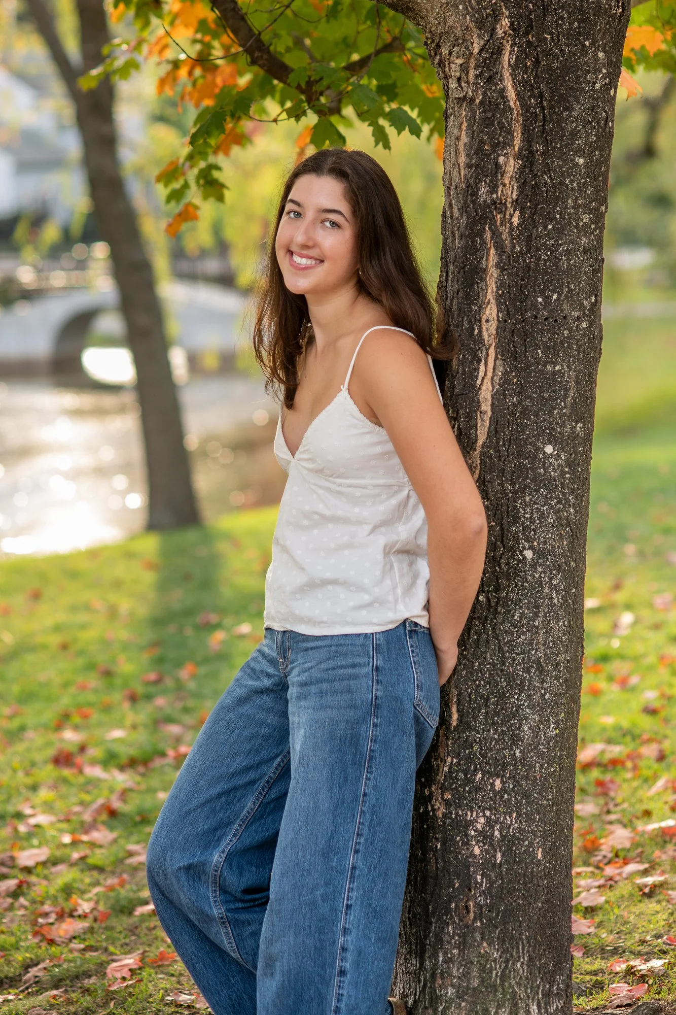 A young woman with long brown hair, wearing a white spaghetti strap top and blue jeans, leaning against a tree in a park during fall, smiling at the camera.
