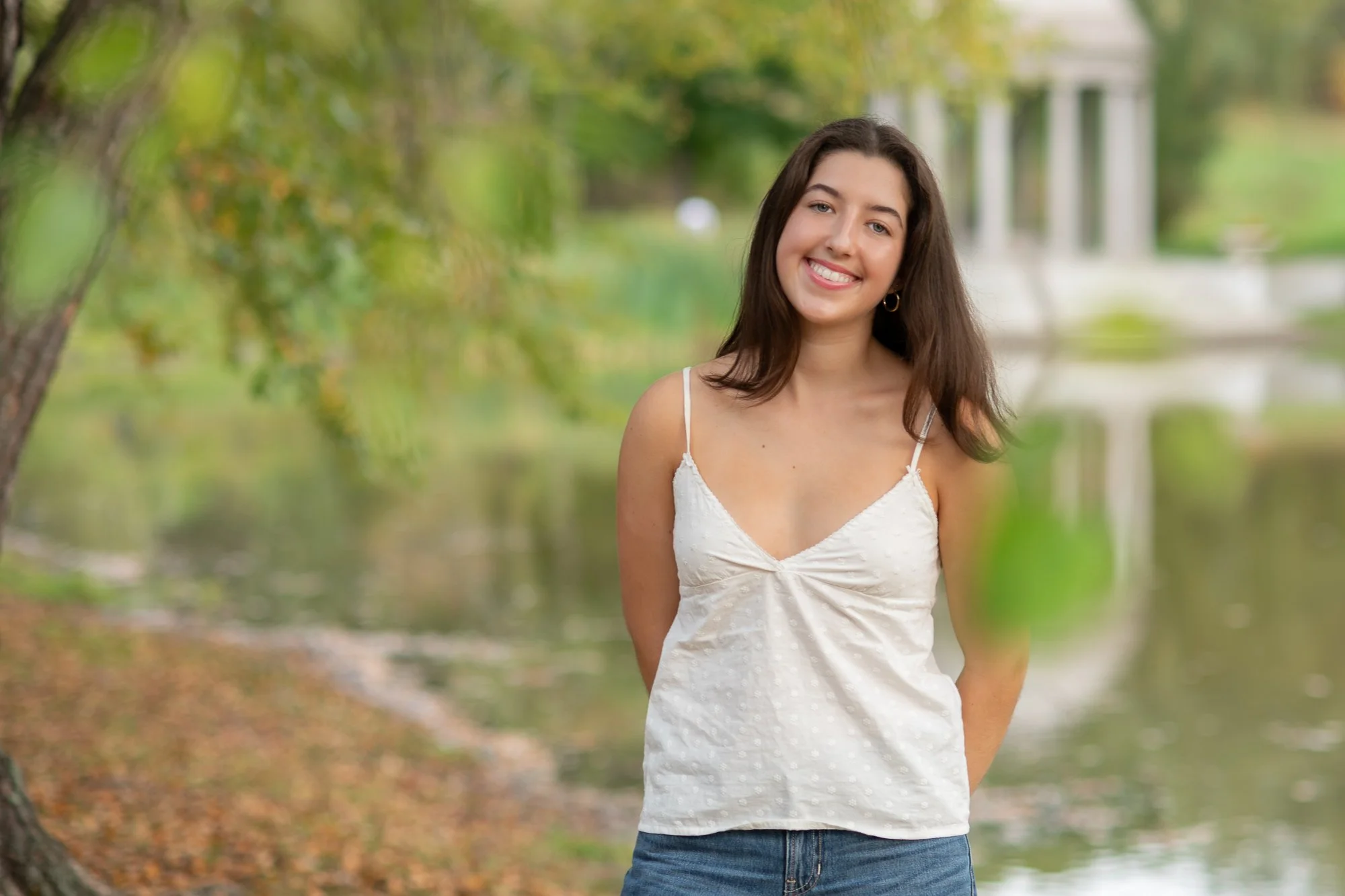 A young woman with brown hair, wearing a white spaghetti strap top and blue jeans, smiling outdoors near a lake with green trees and a white structure in the background.