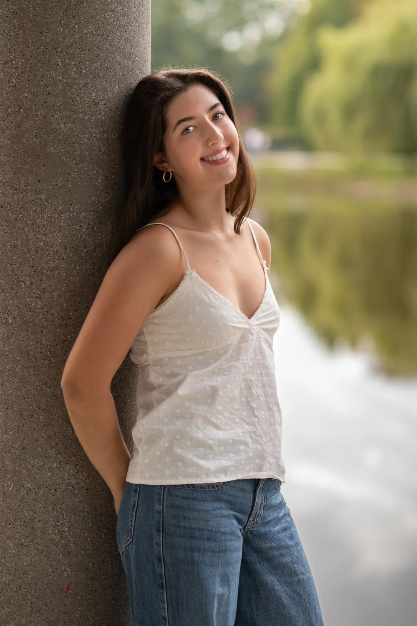 Young woman with long brown hair smiling, leaning against a large concrete column outdoors near a body of water, wearing a white sleeveless top and blue jeans.