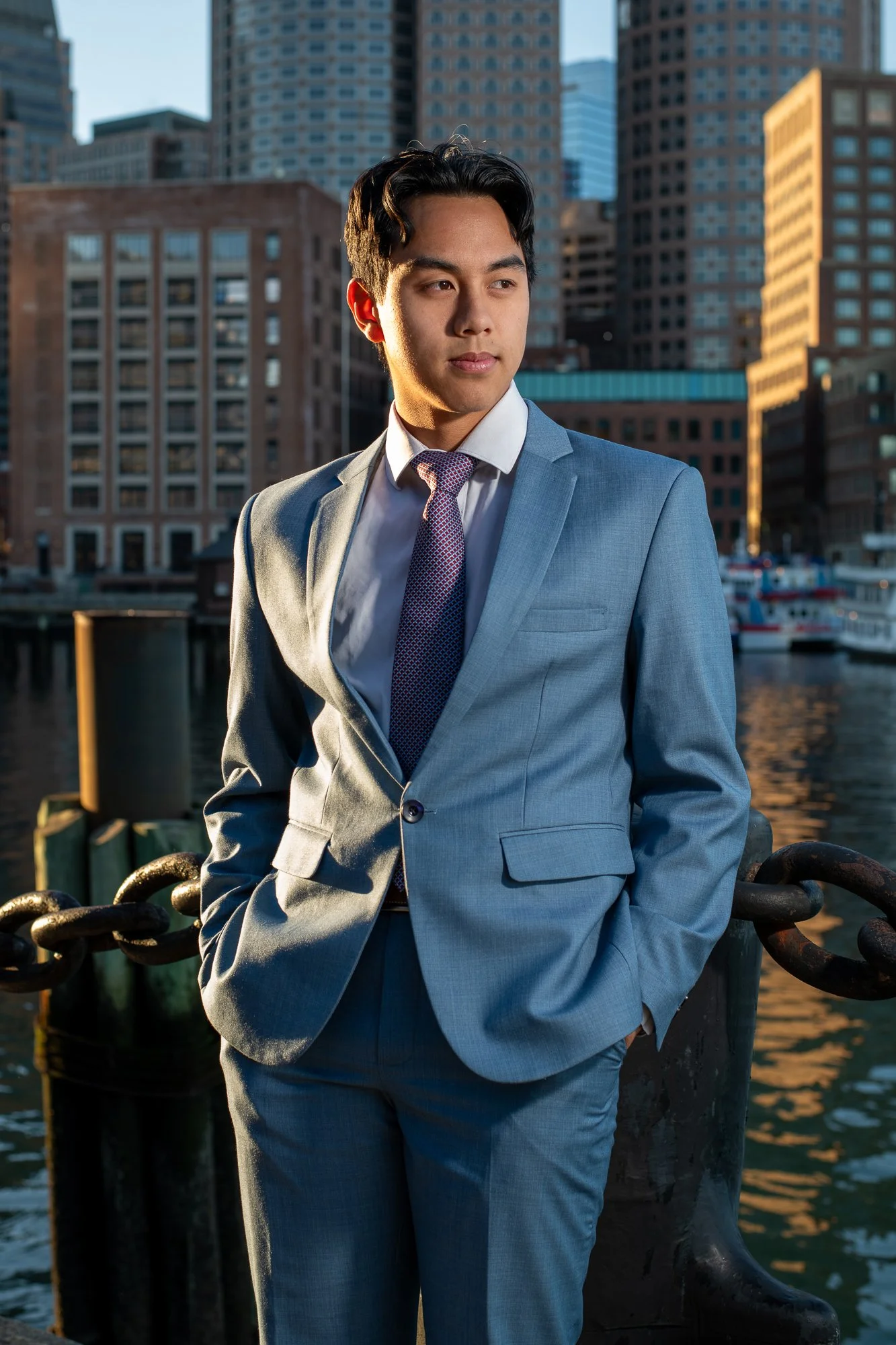 A young man in a light blue suit and tie standing outdoors by a waterfront with city buildings in the background.