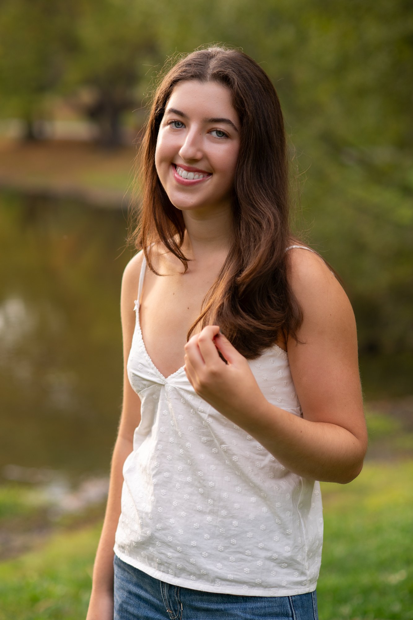 A young woman with long brown hair, light skin, and a white, sleeveless top smiling outdoors next to a body of water, with trees in the background.