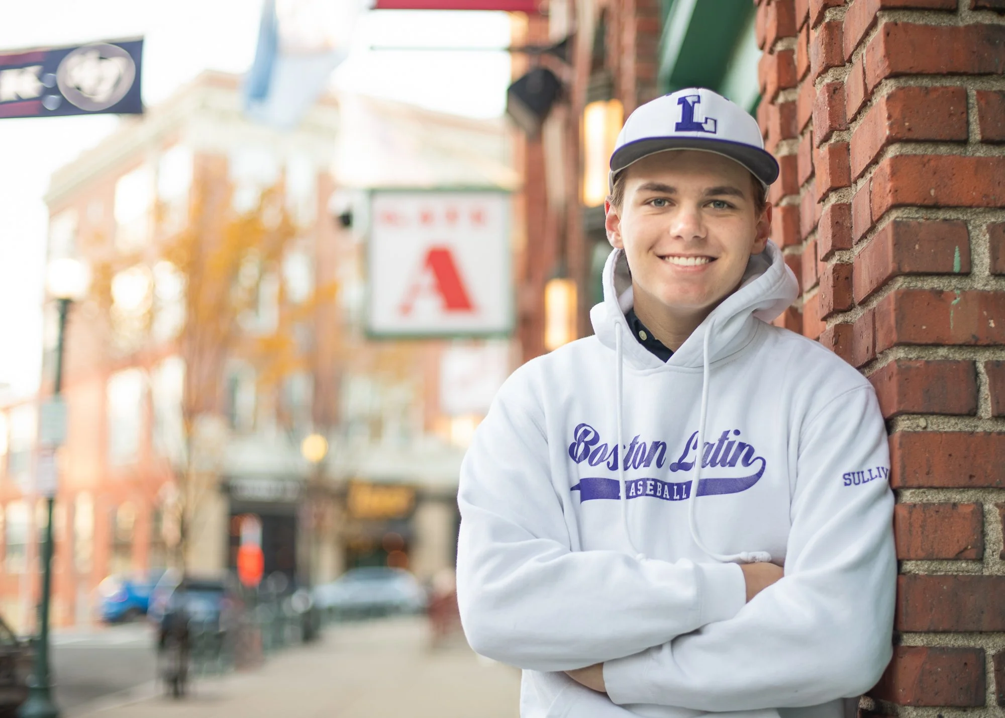 Young man smiling, wearing a baseball cap and hoodie with sports logo, standing against a brick wall outdoors in an urban setting with blurred buildings and street in the background.