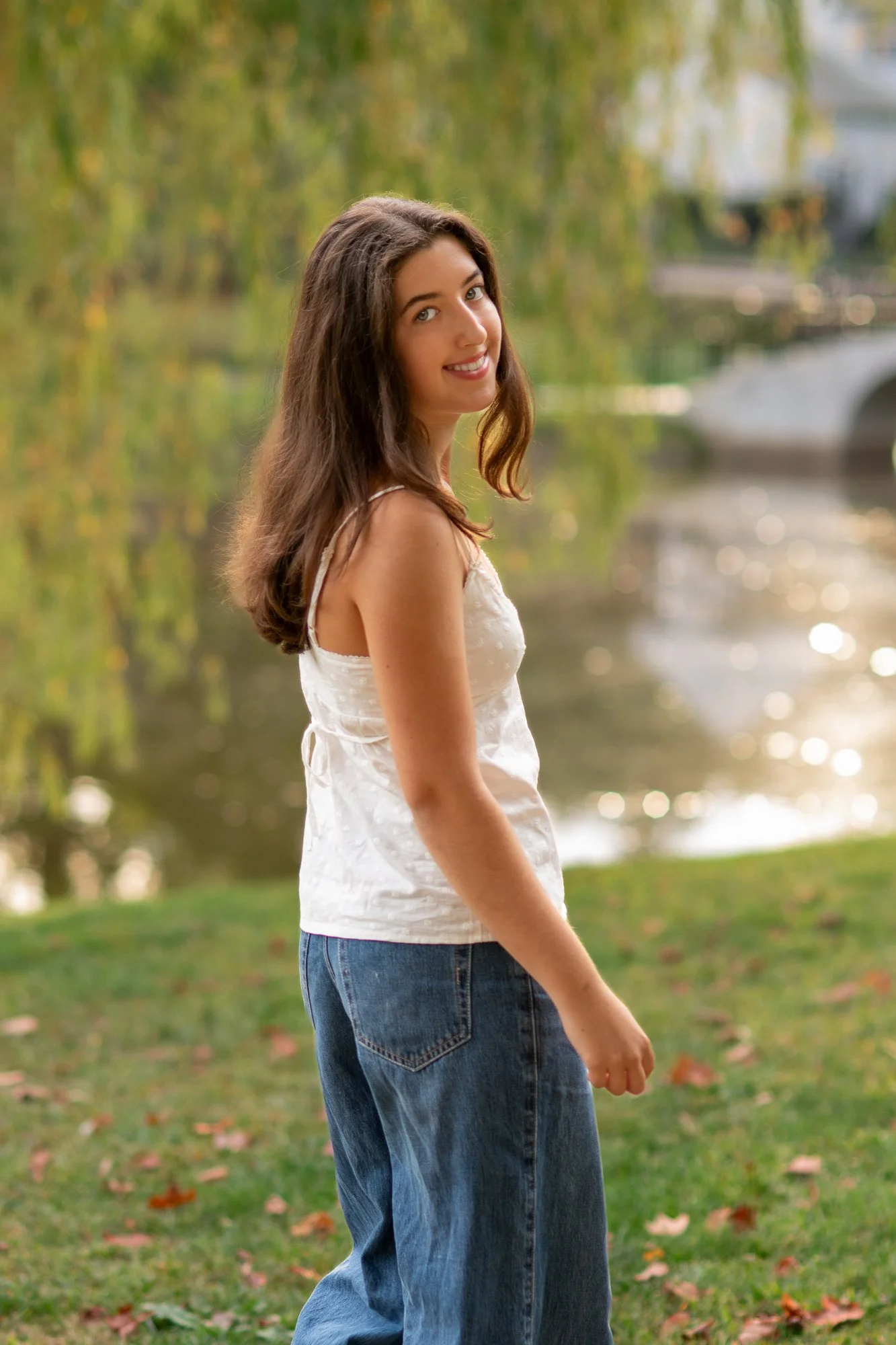 A young woman stands outdoors near a body of water with trees in the background, smiling and looking at the camera.