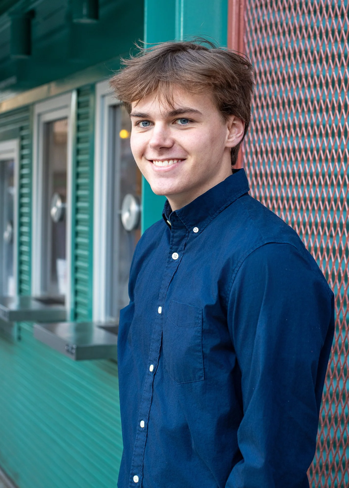 A young man with light brown hair and blue eyes smiling, wearing a dark blue button-up shirt, standing outdoors in front of a building with a teal and red exterior.