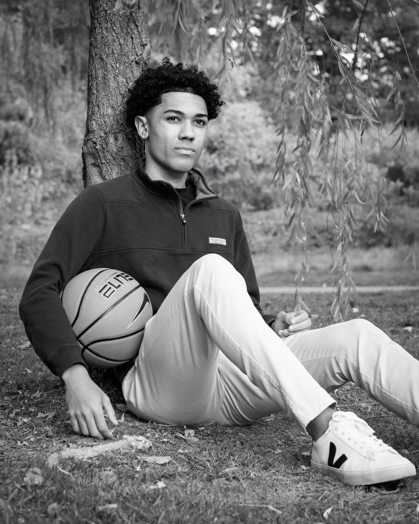 A young person sitting on the grass outdoors, leaning against a tree trunk, holding a basketball, wearing a dark jacket, light pants, and sneakers.