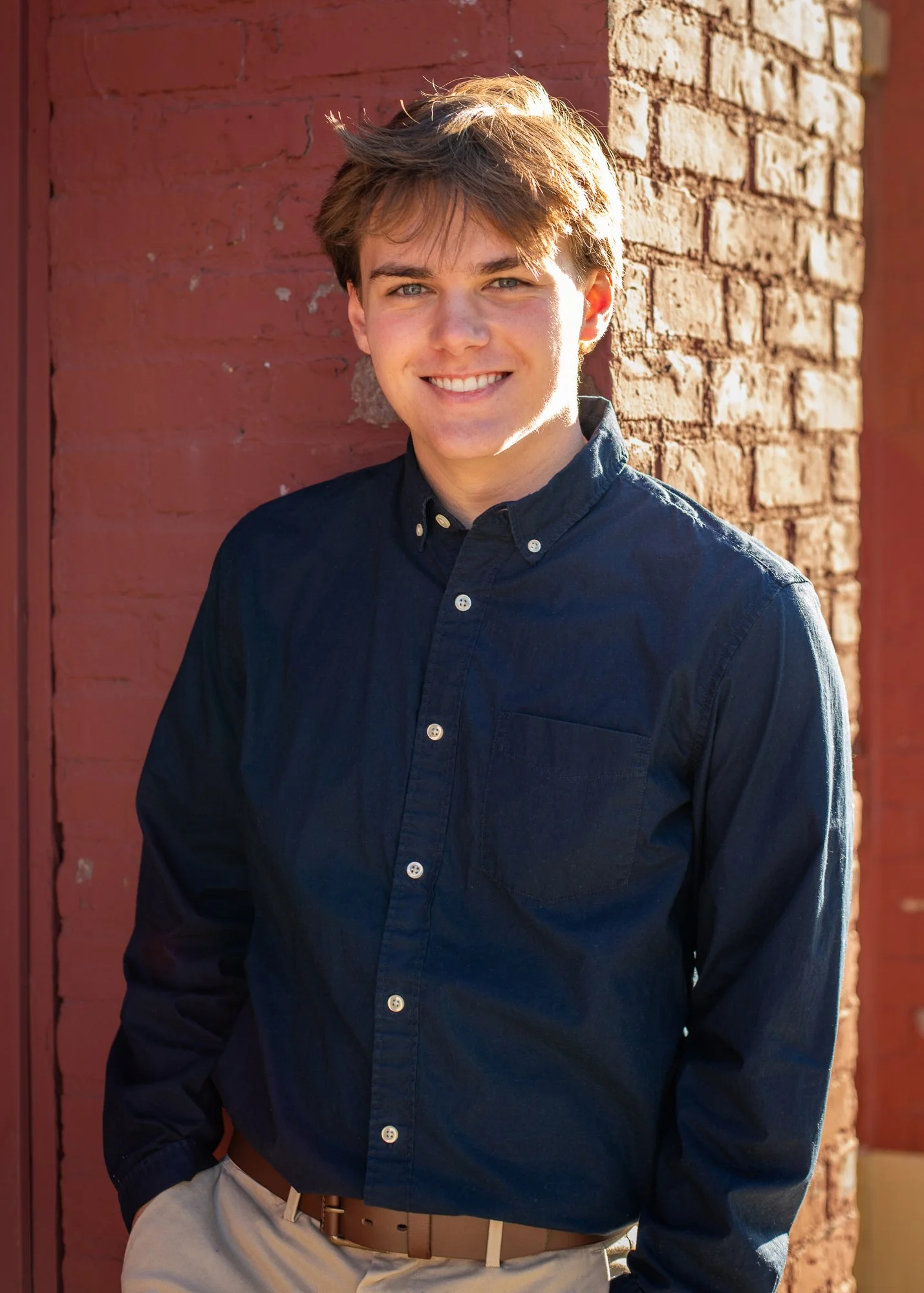 A young man with light brown hair and blue eyes smiling, wearing a dark button-up shirt and light-colored pants, standing outdoors against a red brick wall.