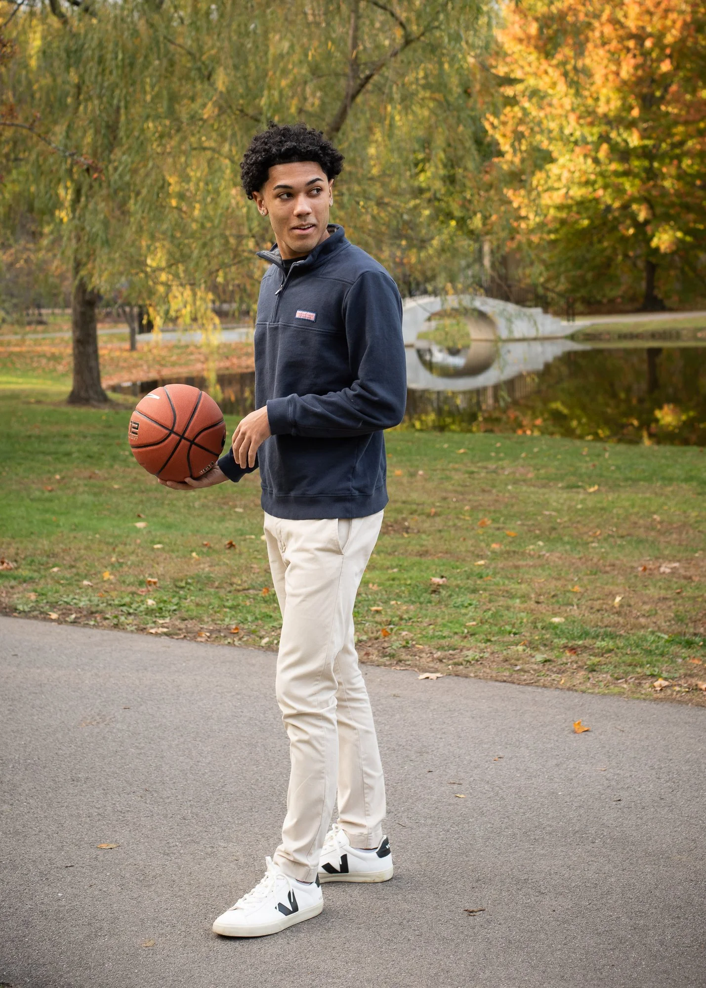 A young man with curly hair standing outdoors on a paved path, holding a basketball, with autumn trees and a pond in the background.