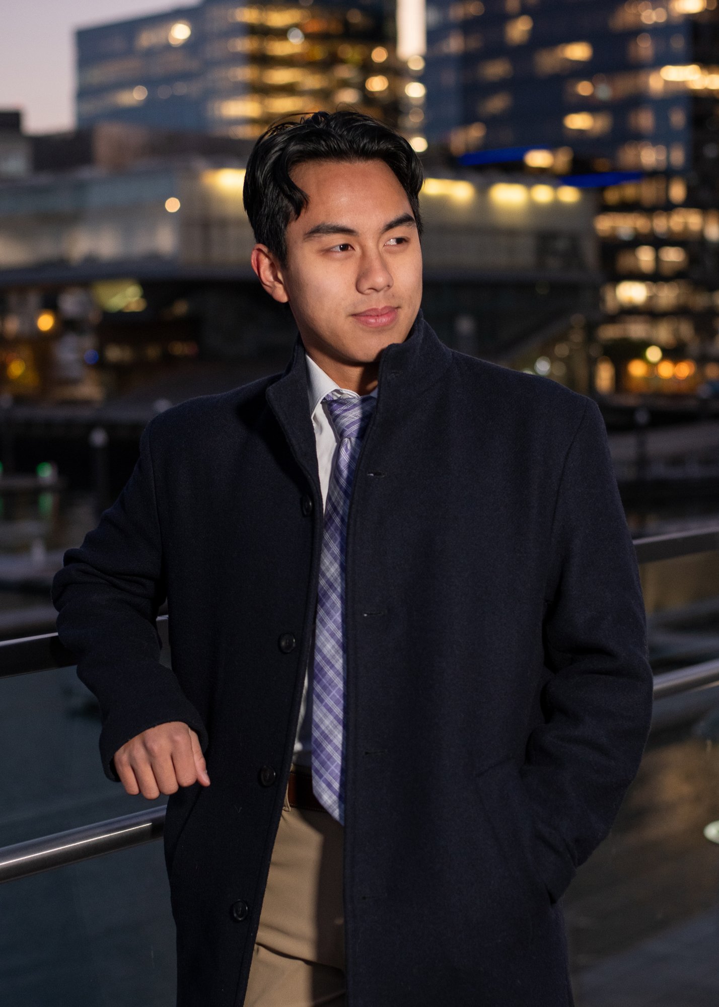 A young man in a dark coat and tie standing outdoors at dusk with a cityscape and marina in the background.