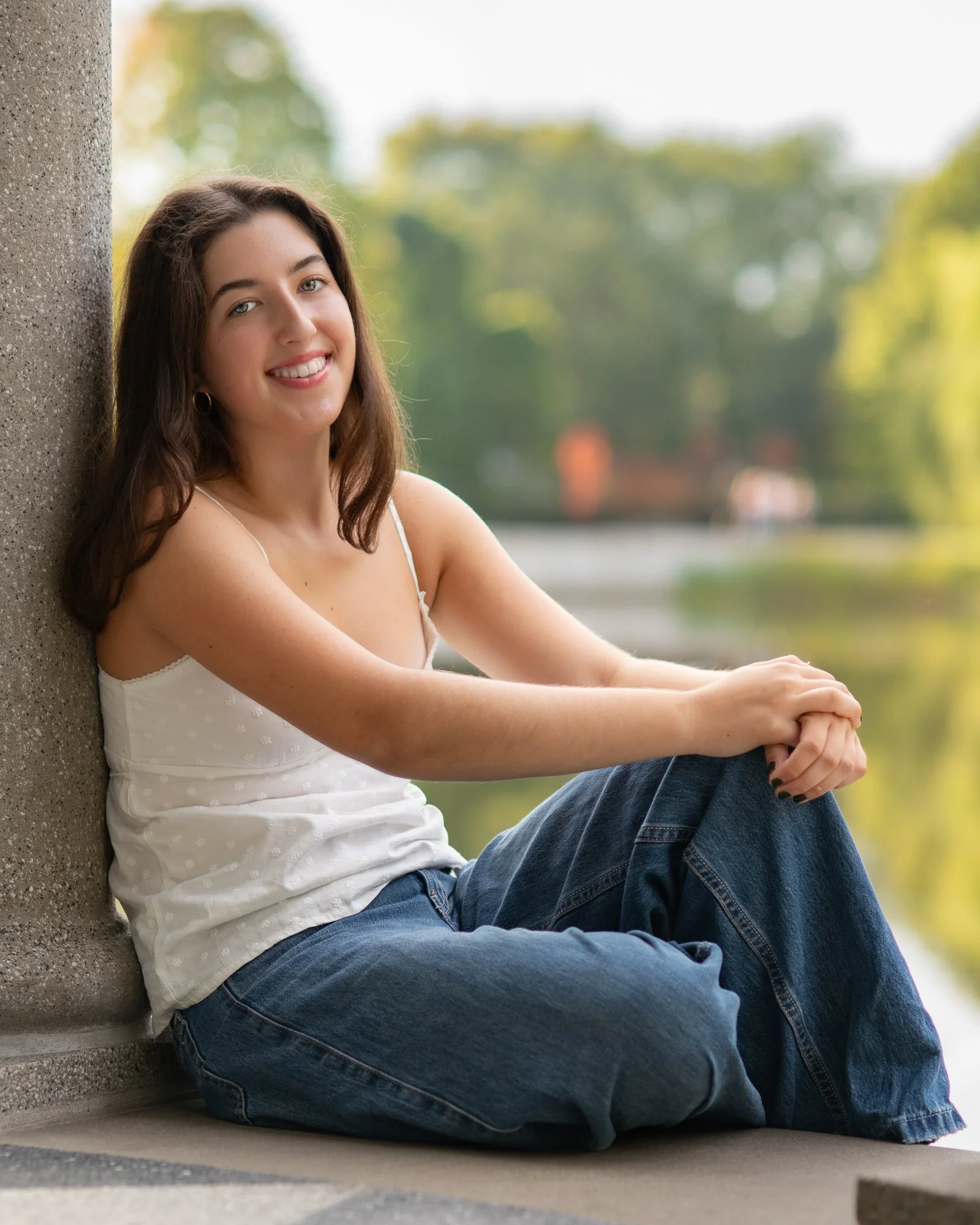 A young woman with brown hair and a white tank top sitting by a concrete wall near a pond, smiling at the camera.