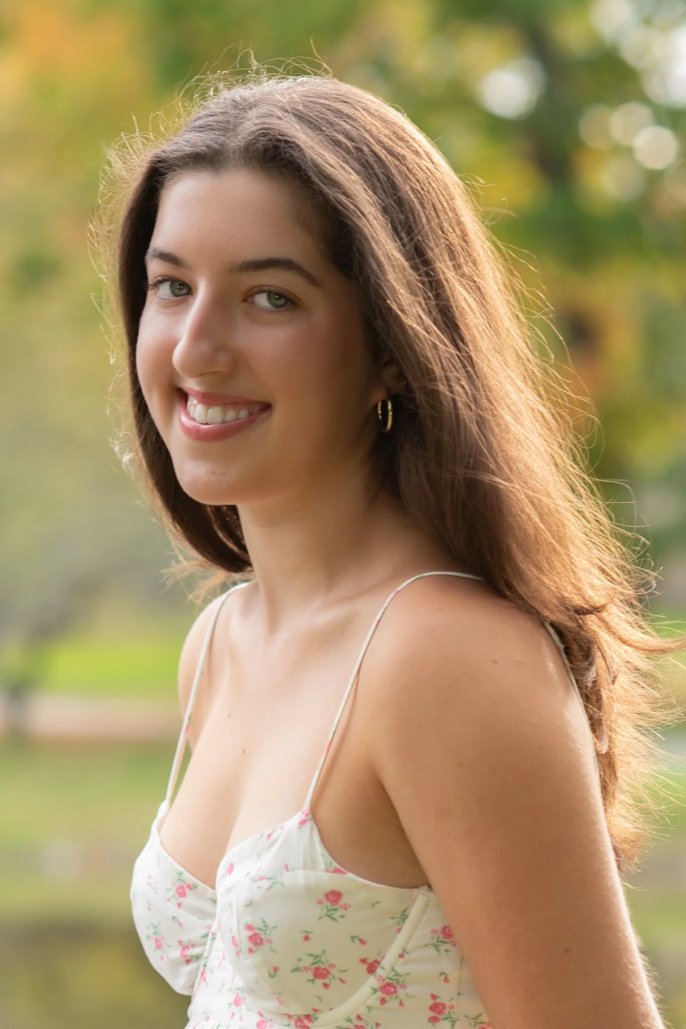 A young woman with long brown hair, green eyes, and a warm smile, standing outdoors in a park with blurred trees and autumn colors in the background.
