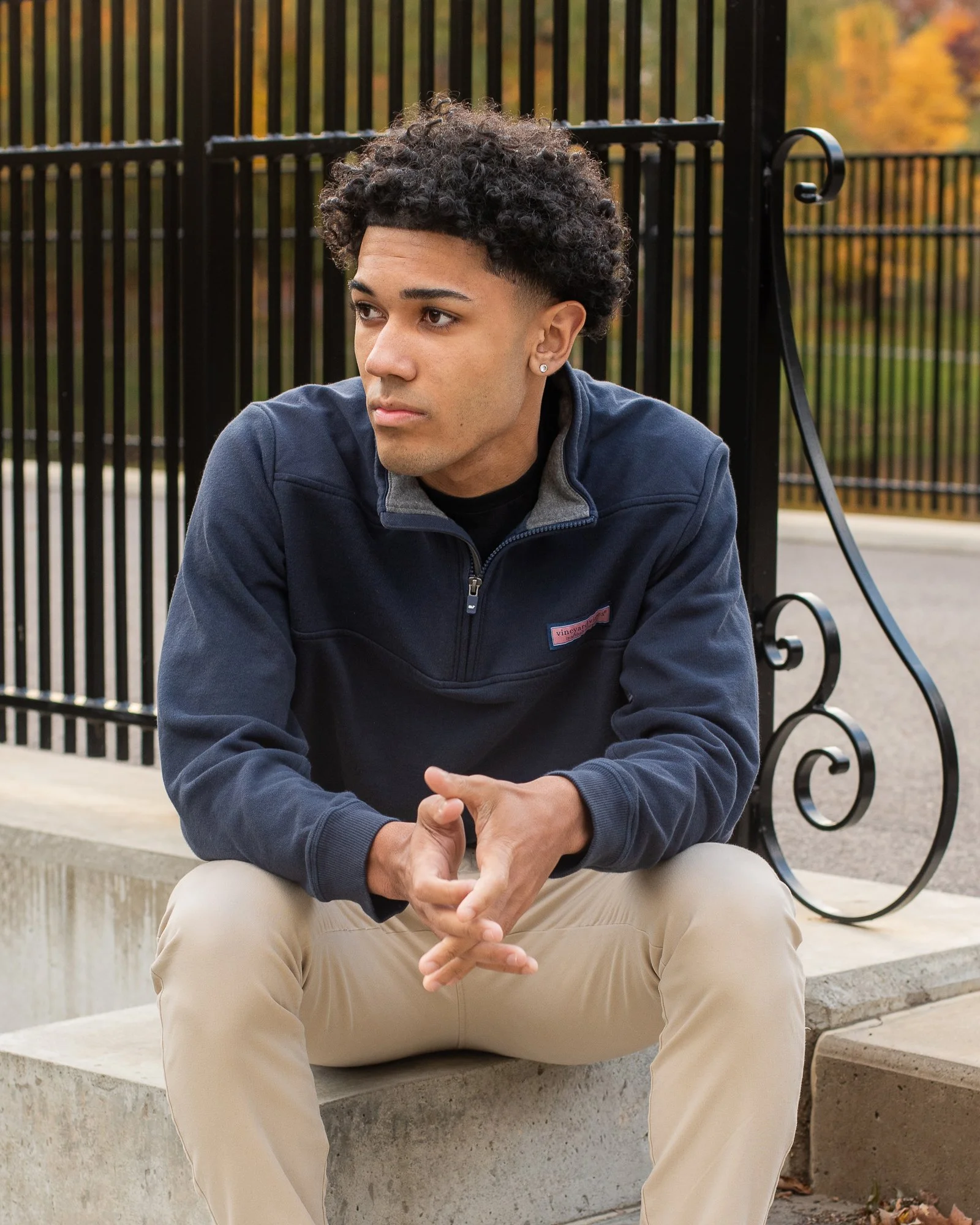 A young man with curly hair, wearing a navy jacket and beige pants, sitting on a concrete ledge in front of a black metal fence outdoors during fall.