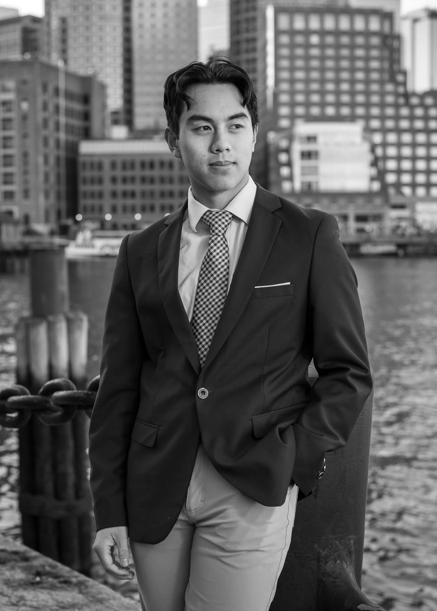 A young man in a business suit standing by a waterfront with city buildings in the background.