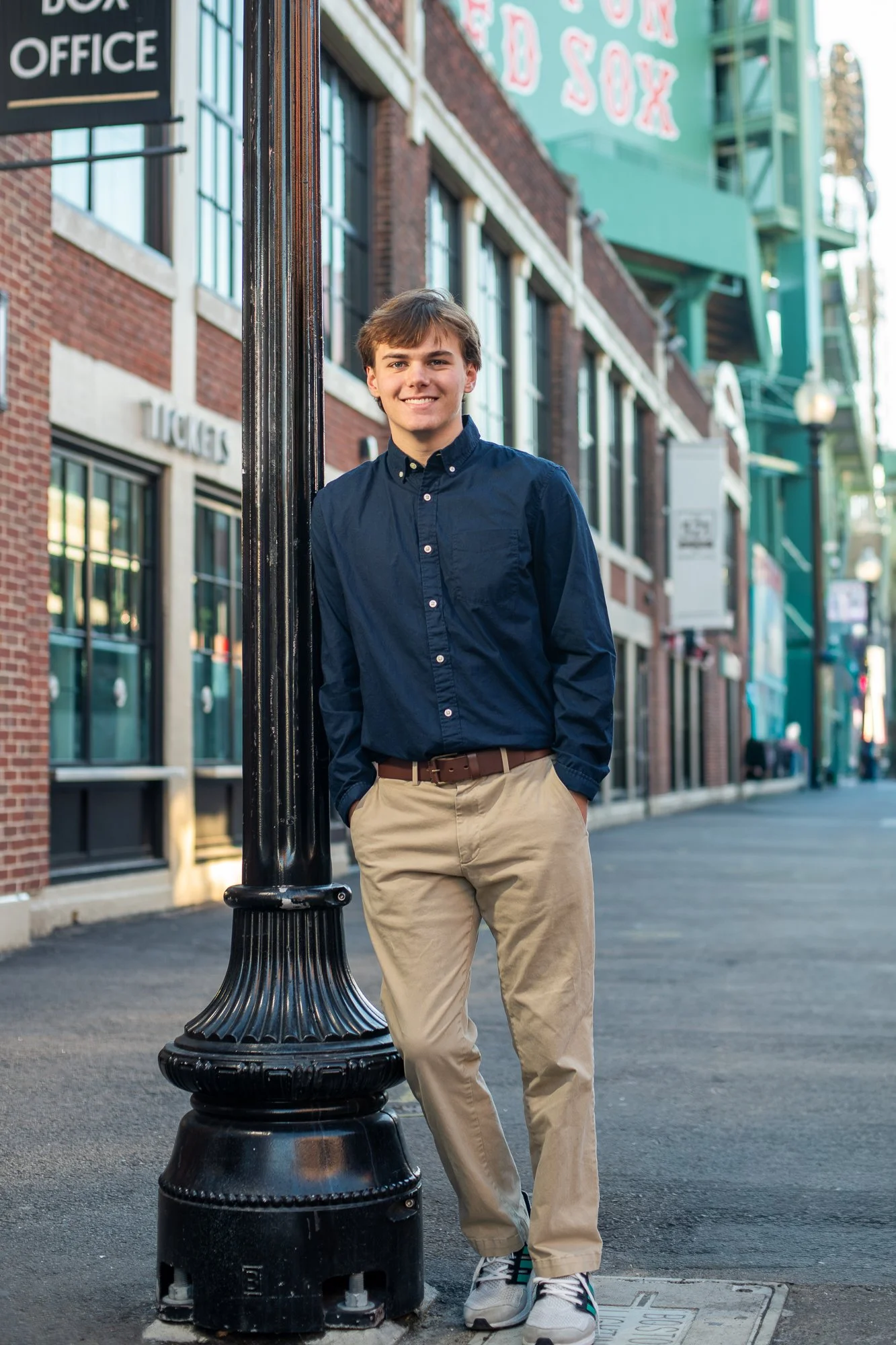 A young man in navy blue shirt and khaki pants leaning against a black lamppost on a city street during daytime.