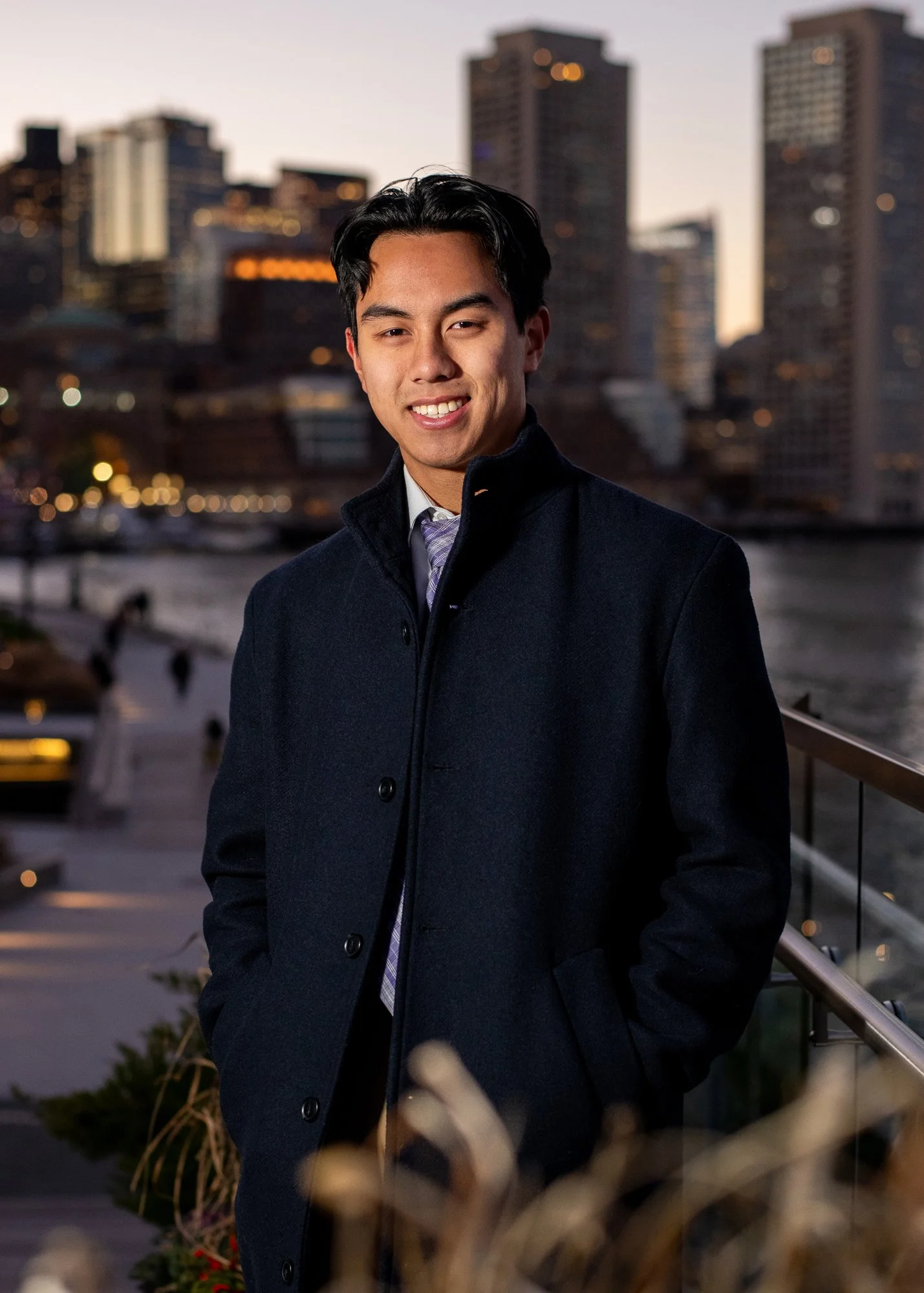A young man in a dark coat and tie standing outdoors against a city skyline at dusk with tall buildings and a river in the background.