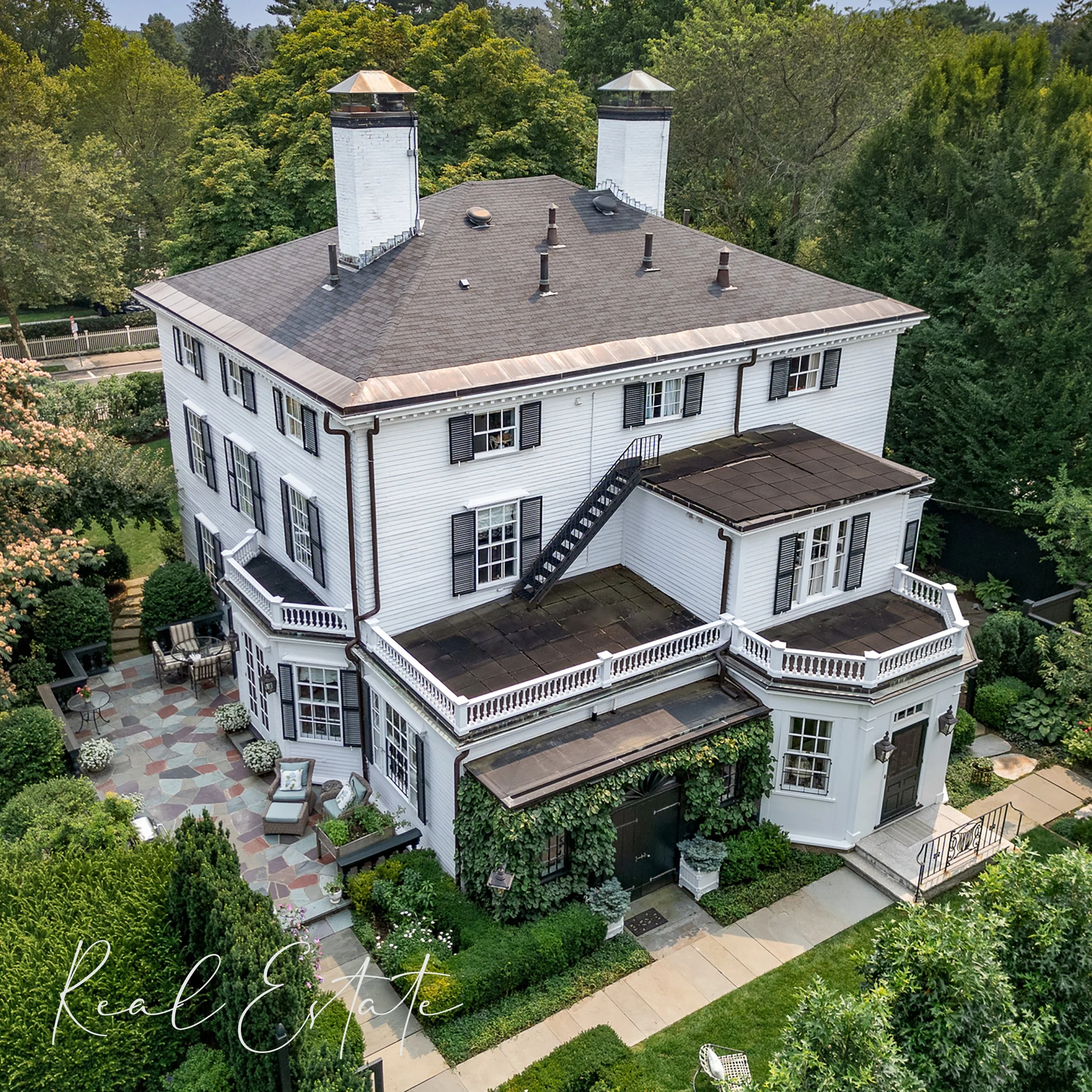 A large, white, multi-story house with black shutters, a gray roof, and a spacious outdoor patio with seating and plants. Surrounded by green trees and grass.