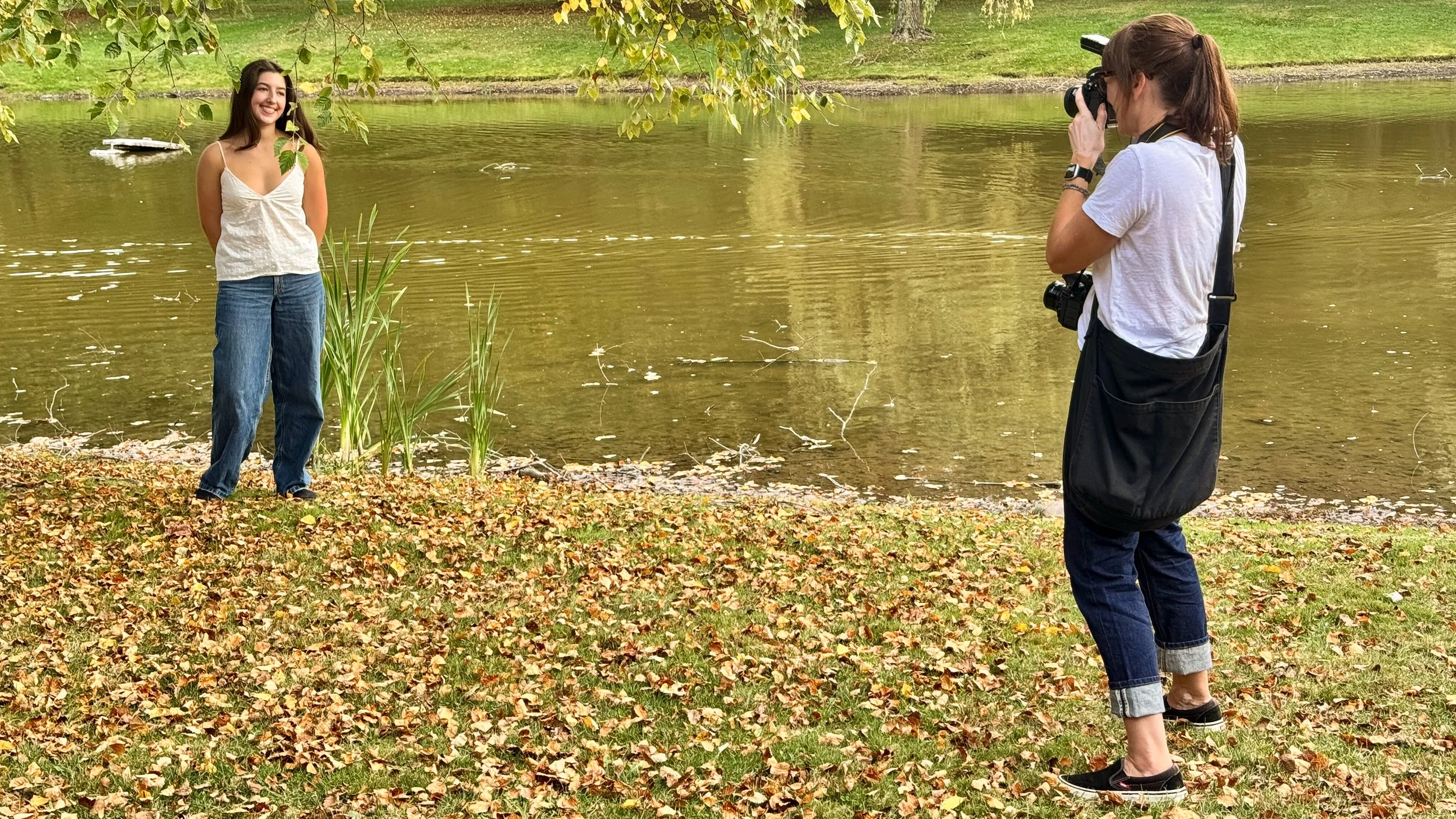 A woman taking a photograph of a smiling woman standing by a lakeside in a park with fallen autumn leaves and trees with green and yellow leaves.