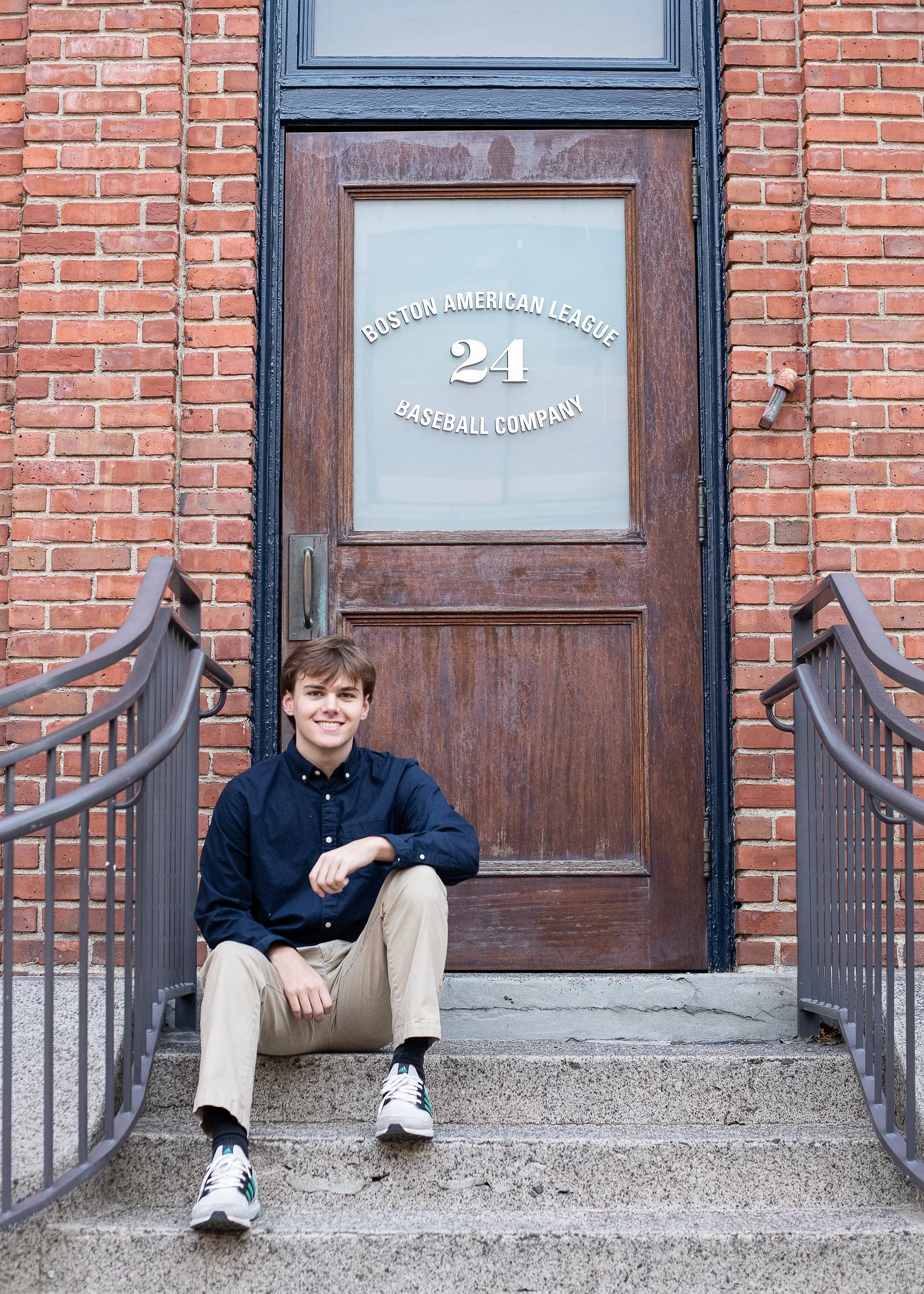 Young man with light brown hair wearing a navy blue shirt, khaki pants, and sneakers sitting on concrete stairs in front of a brown wooden door with a glass window. The door has the text 'Boston American League 24 Baseball Company' on it and is frame