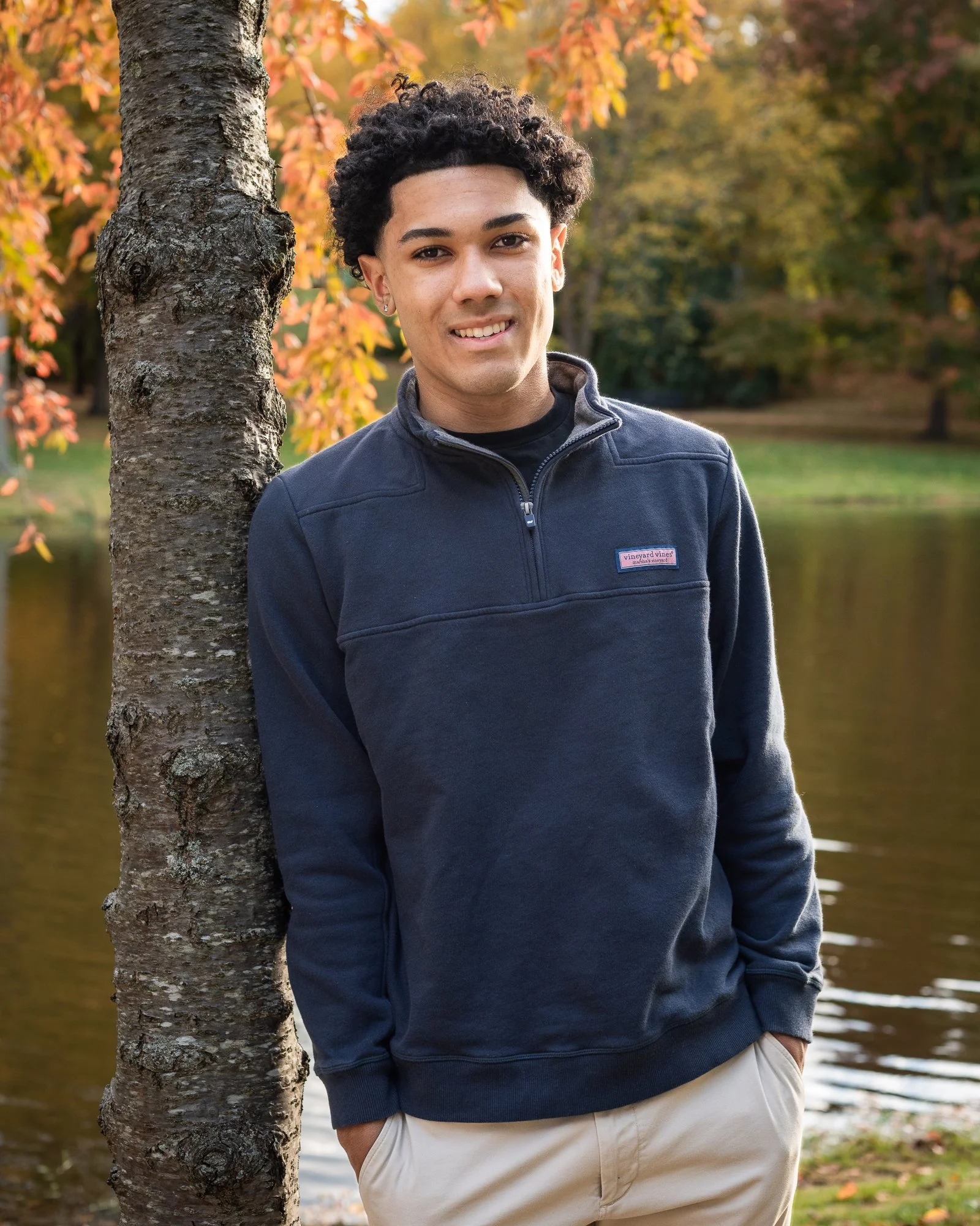 A young man with dark curly hair smiling, wearing a dark blue sweatshirt, standing outdoors near a tree by a river with autumn trees in the background.