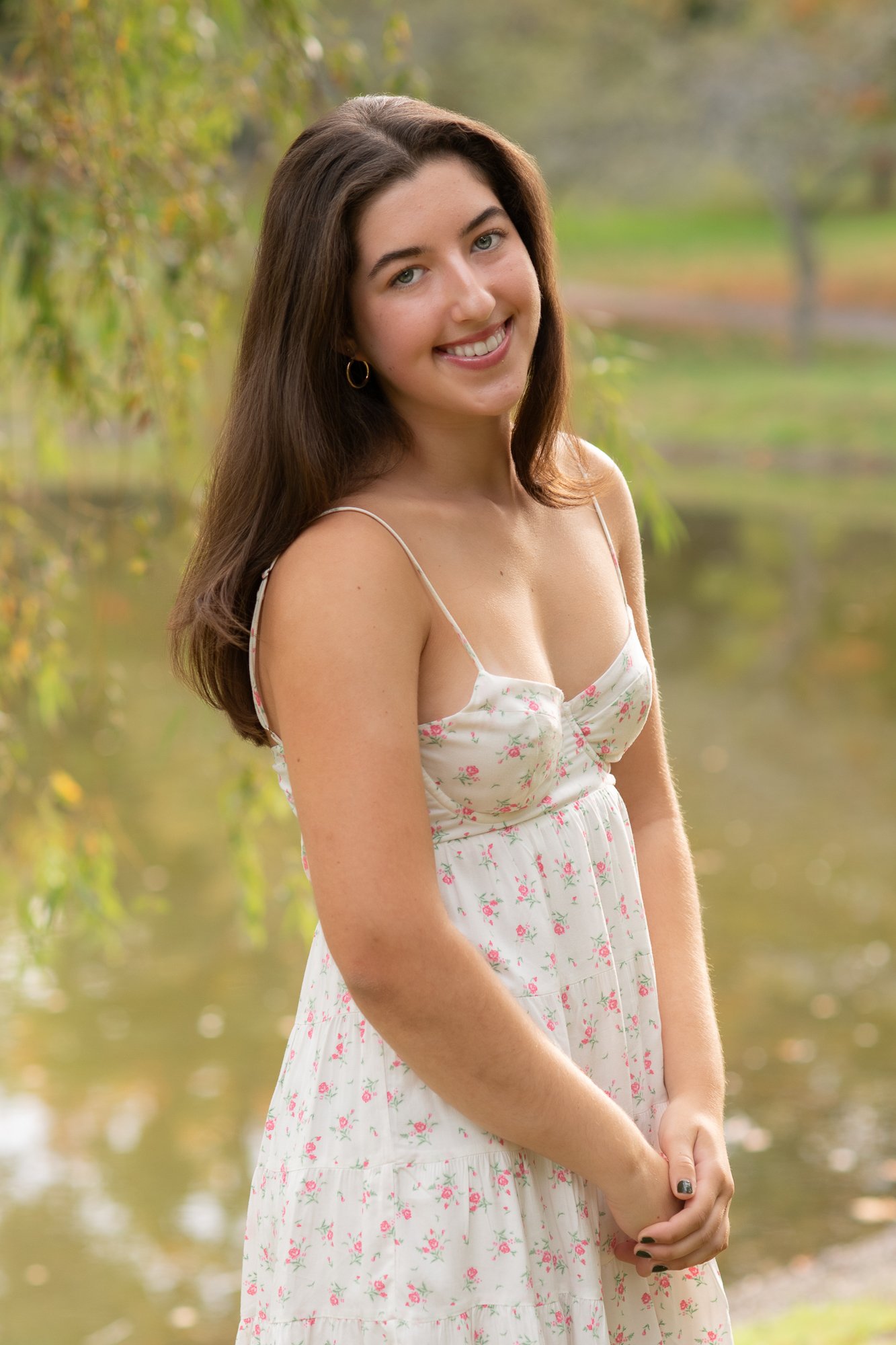 A young woman with long brown hair, wearing a white floral dress with spaghetti straps, stands outdoors near a body of water with trees and greenery in the background, smiling at the camera.