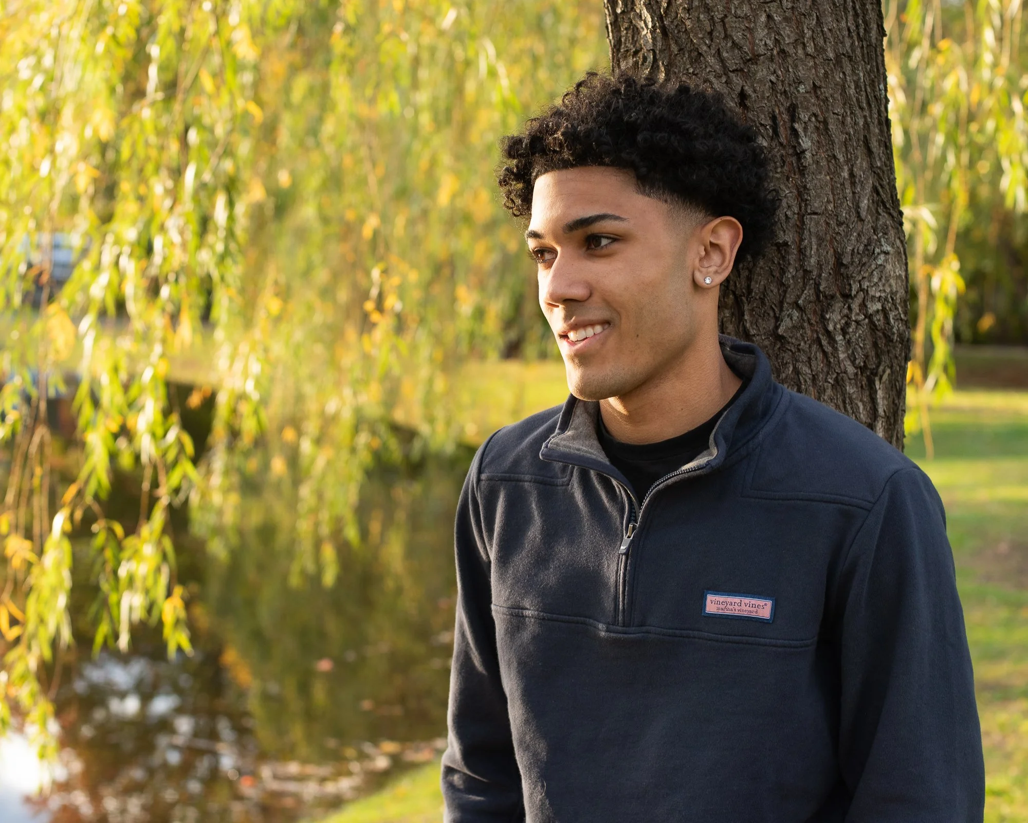A young man with curly black hair, earrings, and a navy vineyard vines jacket, standing by a tree in a park with yellow-green autumn foliage.