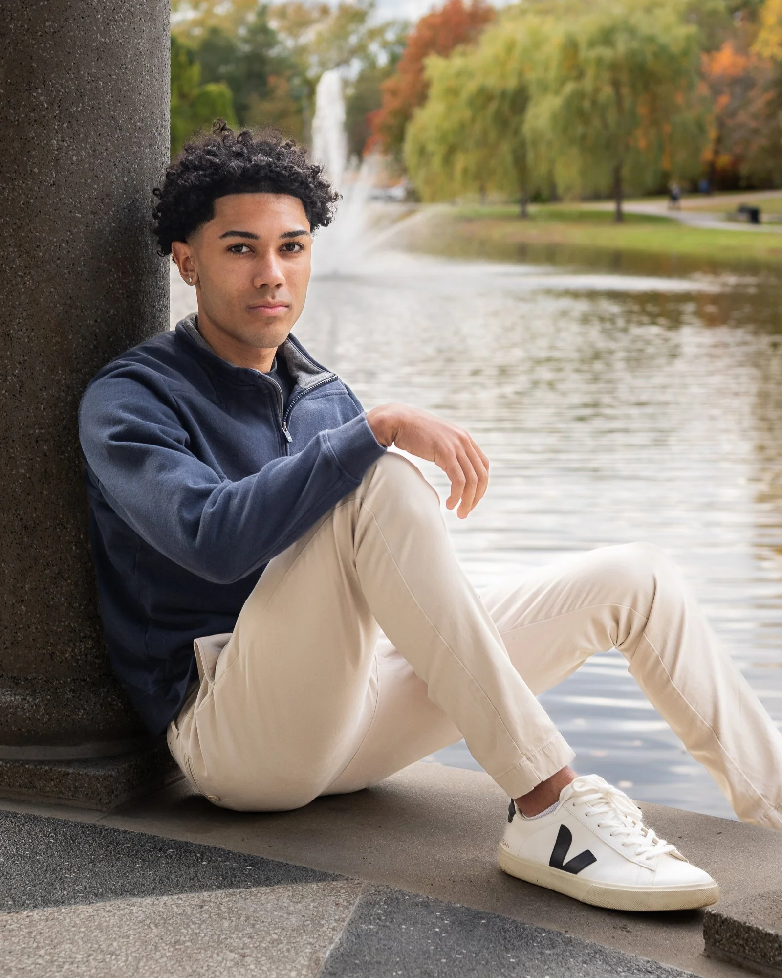 A young man with curly dark hair sitting cross-legged by a body of water, leaning against a concrete pillar, with trees and a fountain in the background during fall.