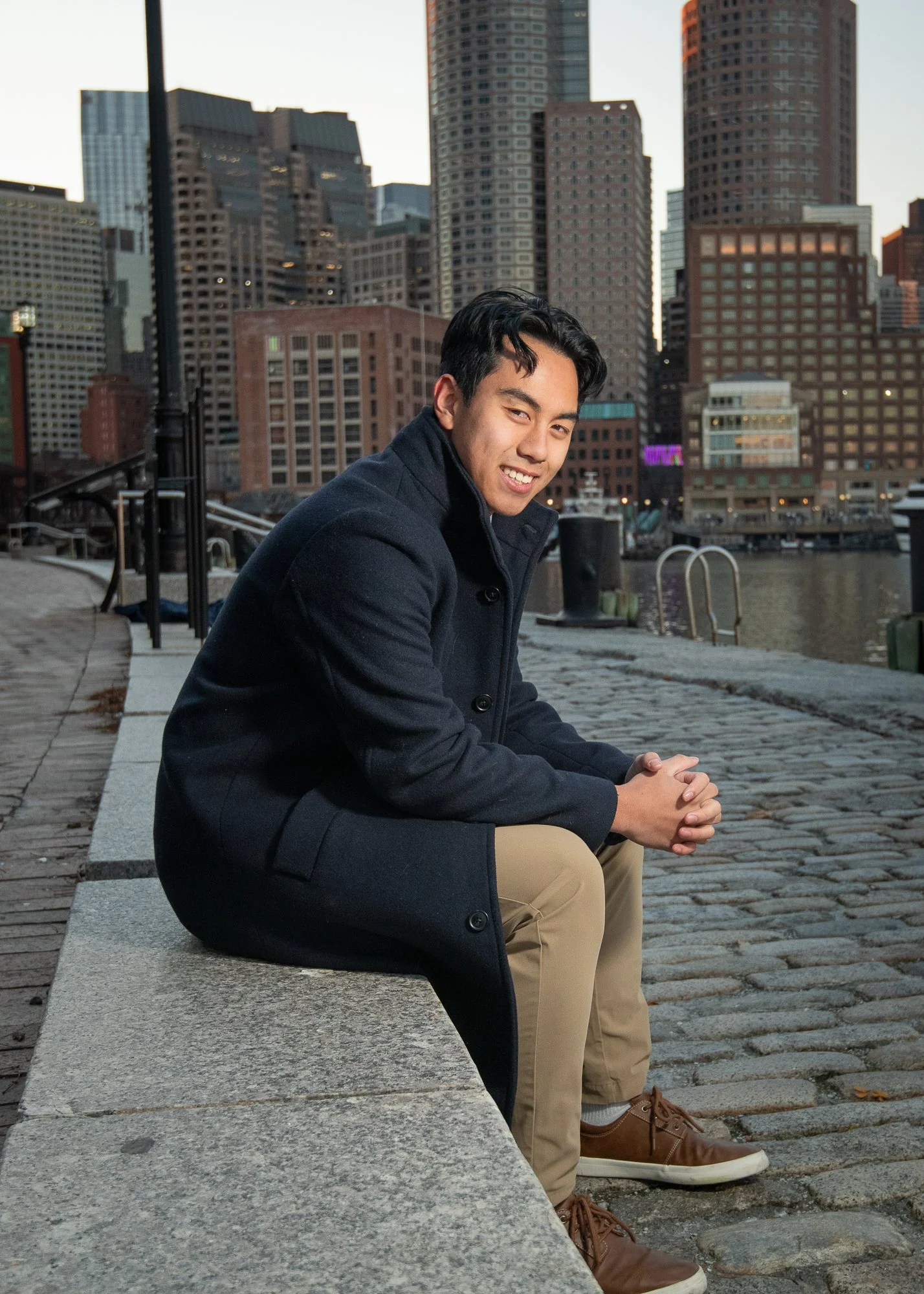 A young man sitting on a bench by a river in a city at sunset, smiling and wearing a dark coat and khaki pants.