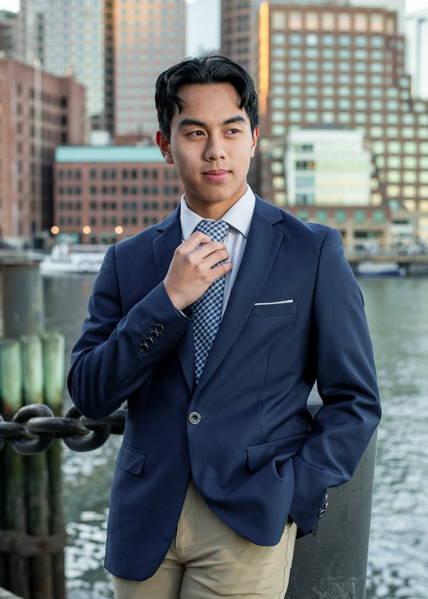 A young man in a navy blue blazer, white shirt, checkered tie, and beige pants standing outdoors by the water in an urban setting with tall buildings in the background.
