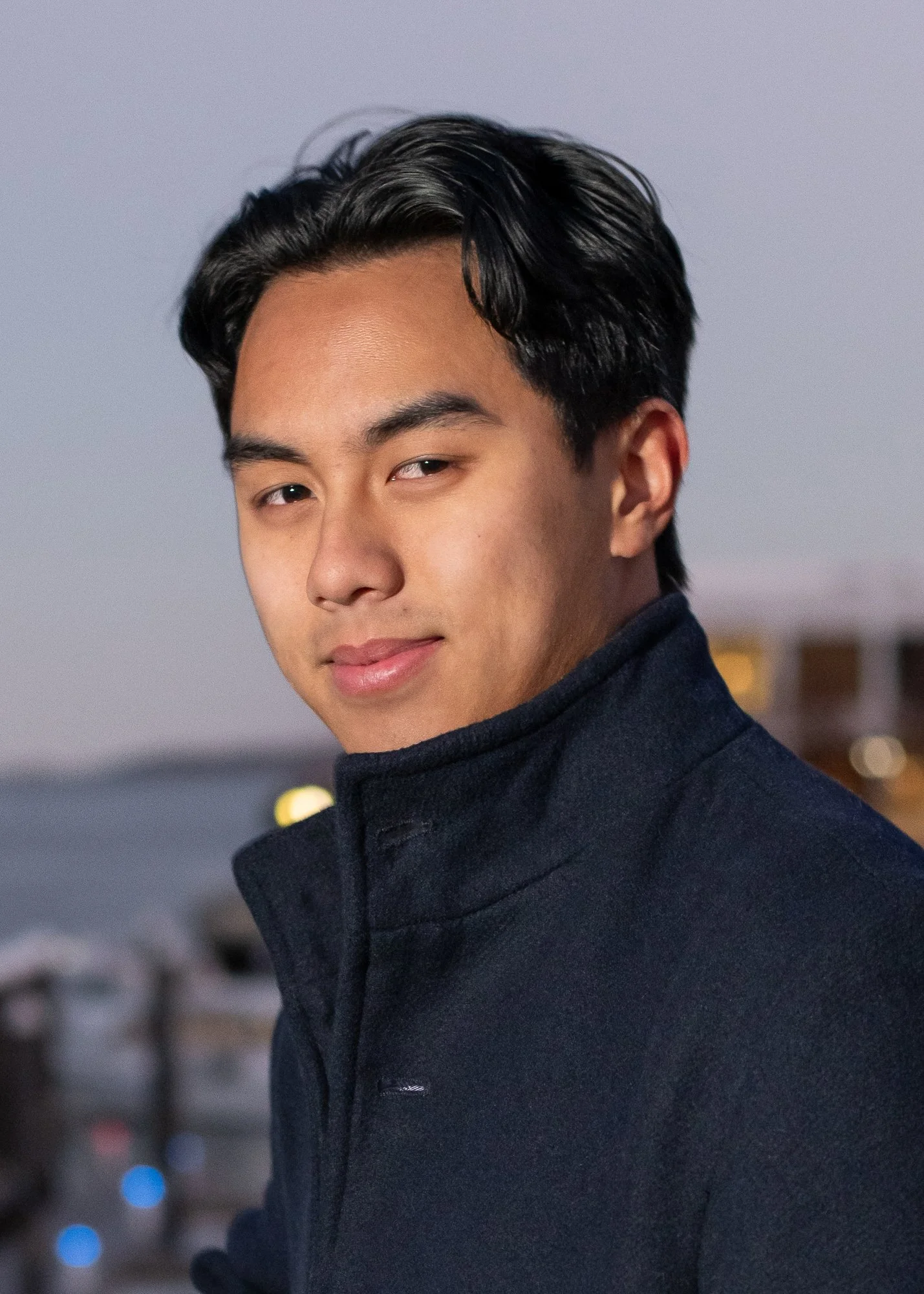 A young man with short black hair and a slight smile, wearing a dark jacket, standing outdoors during dusk with a blurred cityscape in the background.