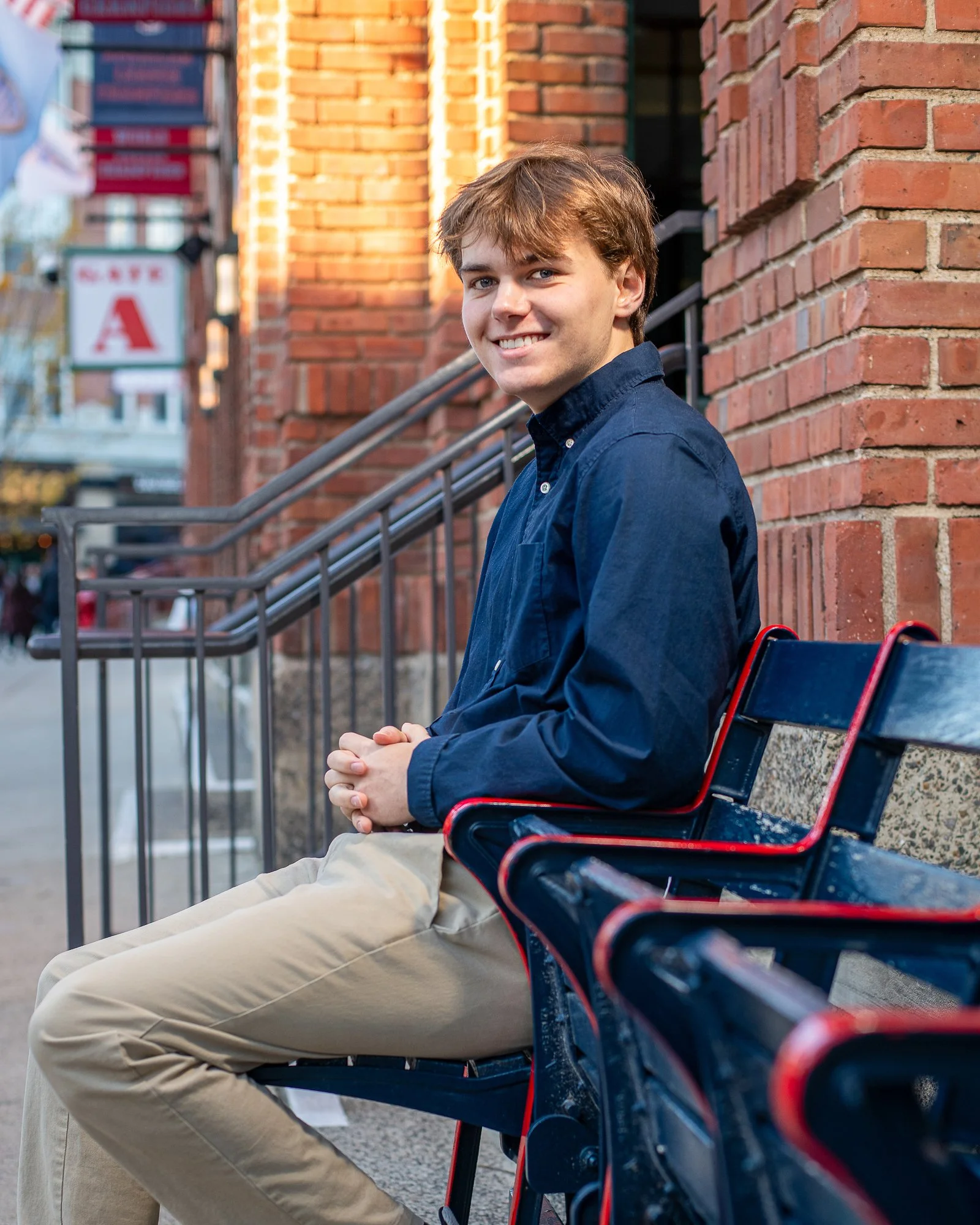 A young man with light brown hair, wearing a dark blue shirt and beige pants, sitting on a city bench beside a brick building, smiling at the camera.