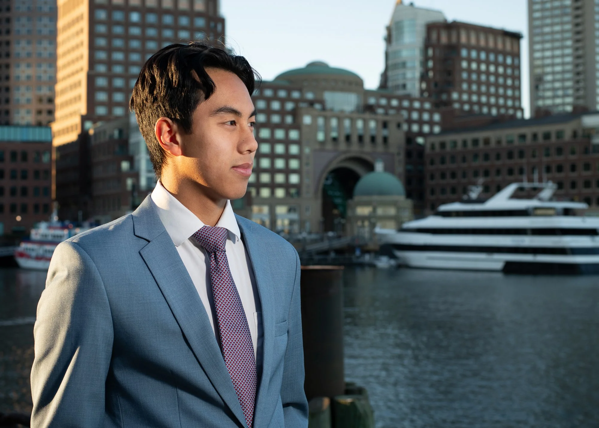 A young man in a gray suit and tie standing outdoors near a harbor with yachts and tall buildings in the background.