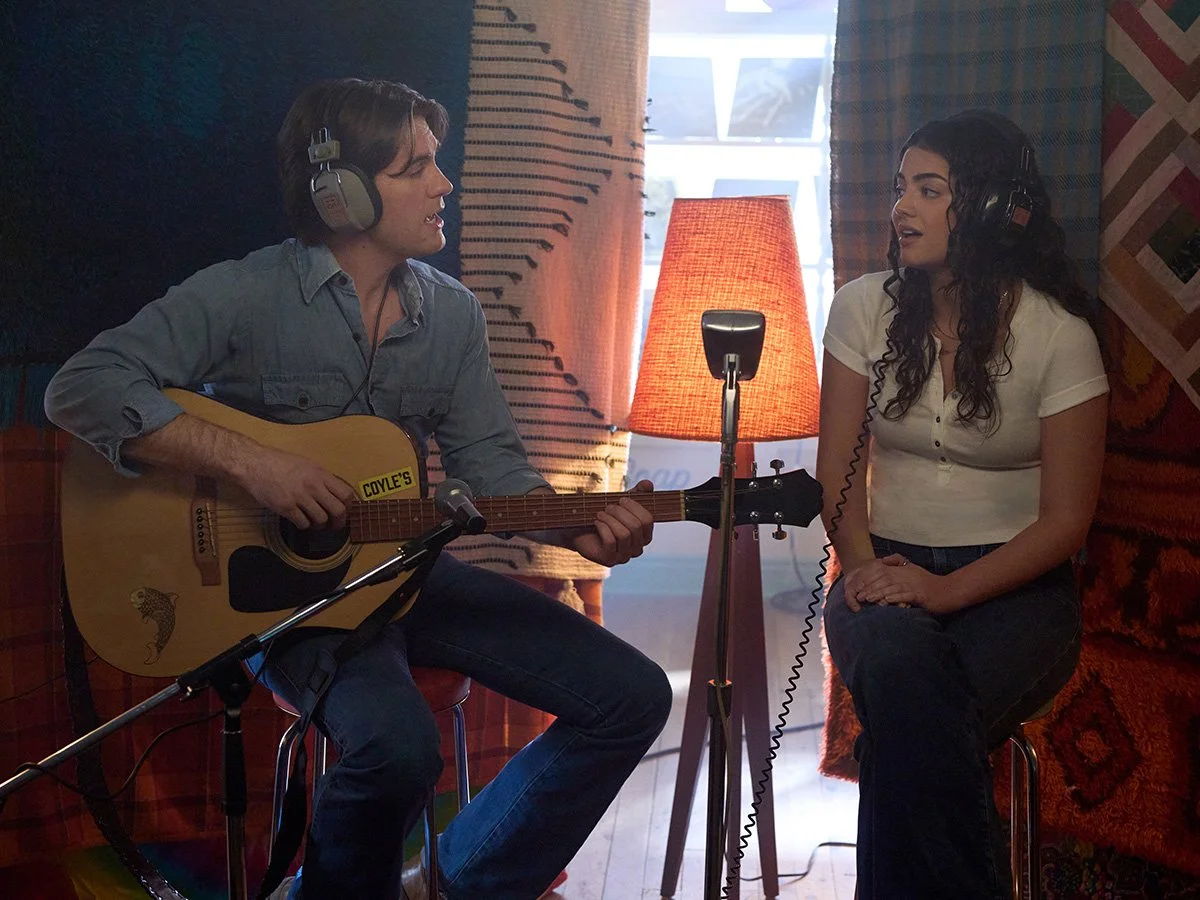 A man and a woman in a cozy room with woven tapestry wall hangings, sitting in front of a window with sunlight. The man plays an acoustic guitar and wears headphones, while the woman listens and sings into a microphone, also wearing headphones.