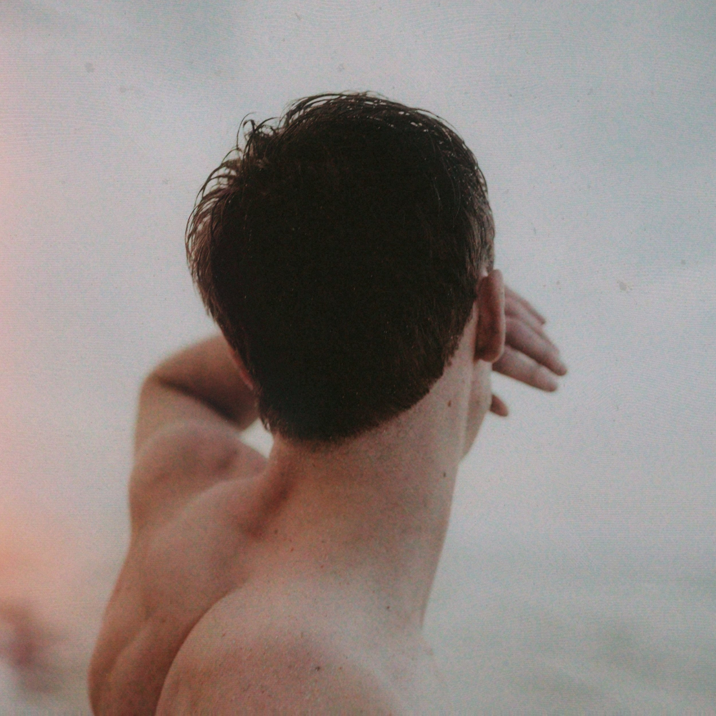 From behind view of a shirtless man touching their forehead with their left hand, with short dark hair and fair skin, against a plain background.