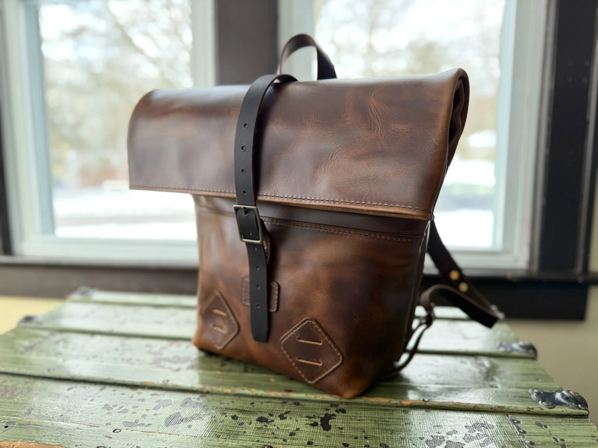 A brown leather backpack with a buckle strap, placed on a rustic wooden surface near a window, with a blurred outdoor background.