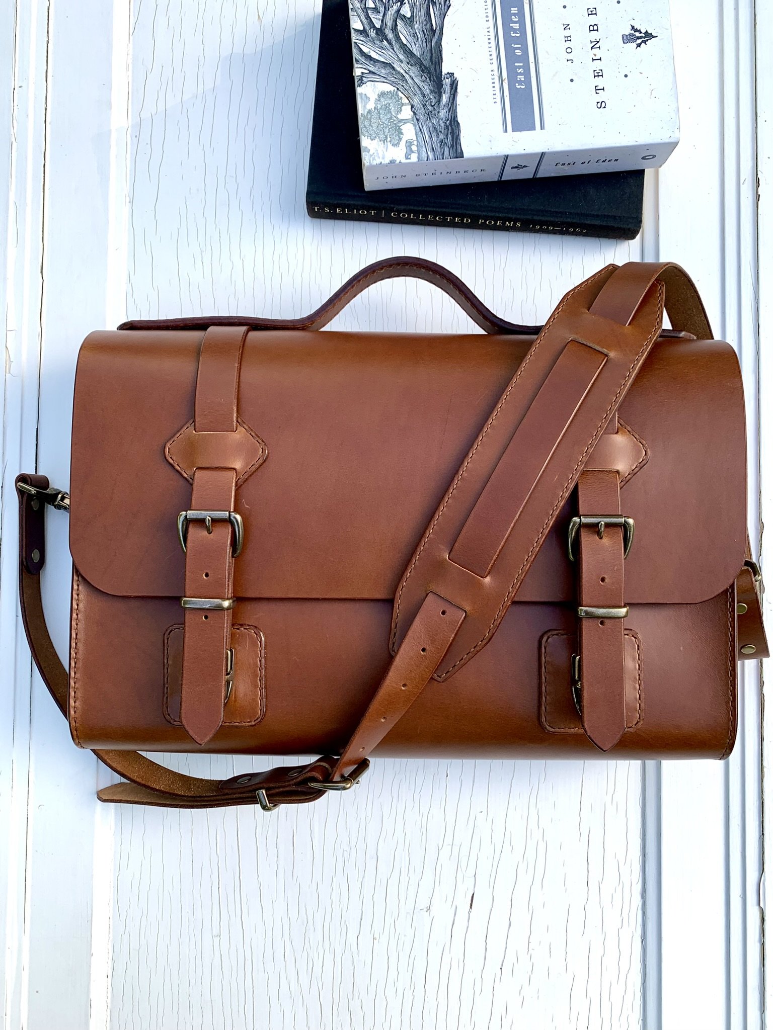 Top view of a brown leather briefcase on a white wooden surface, with a book and a journal behind it.