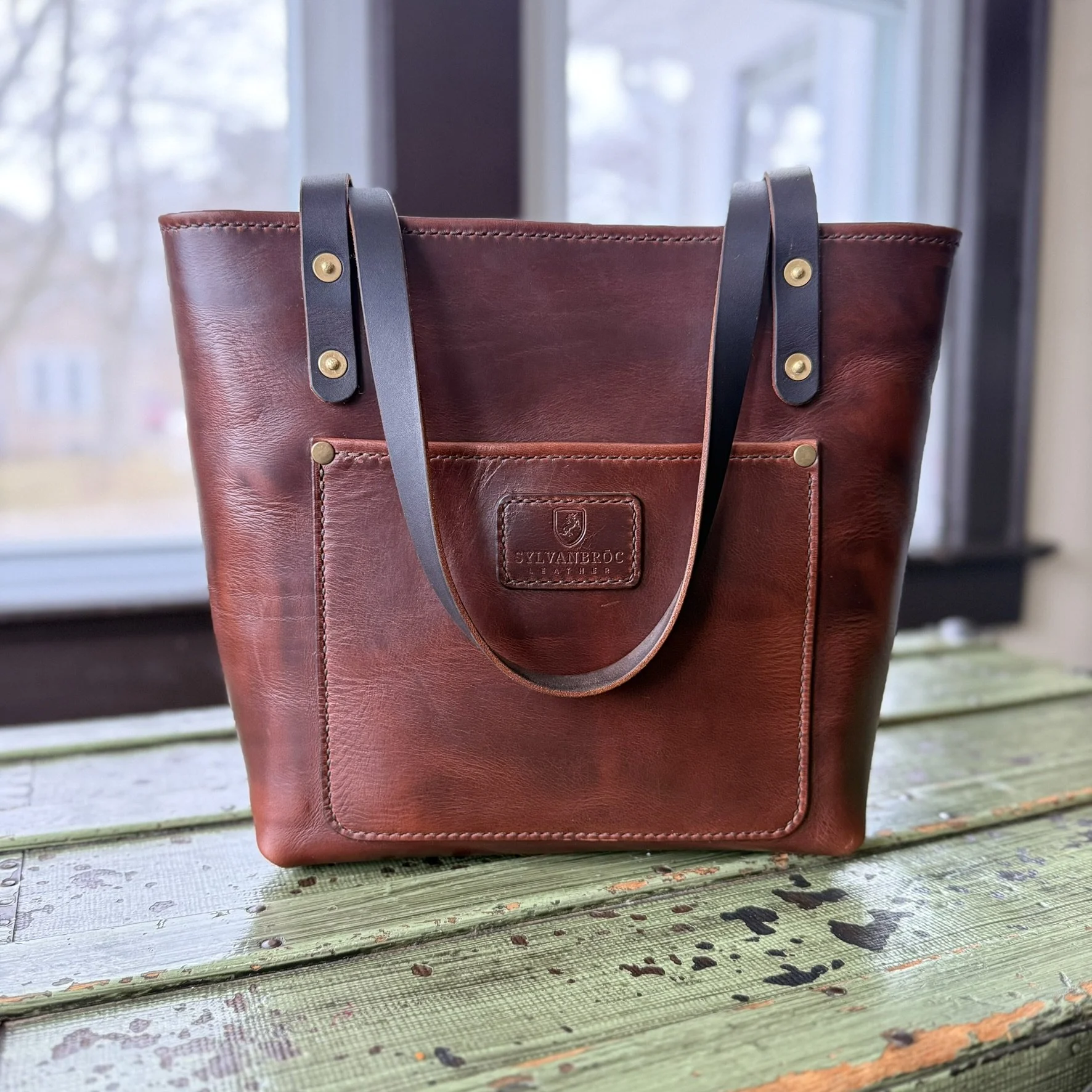 Brown leather tote bag with dark leather straps and gold hardware, placed on a weathered green wooden surface, with a blurred window in the background.