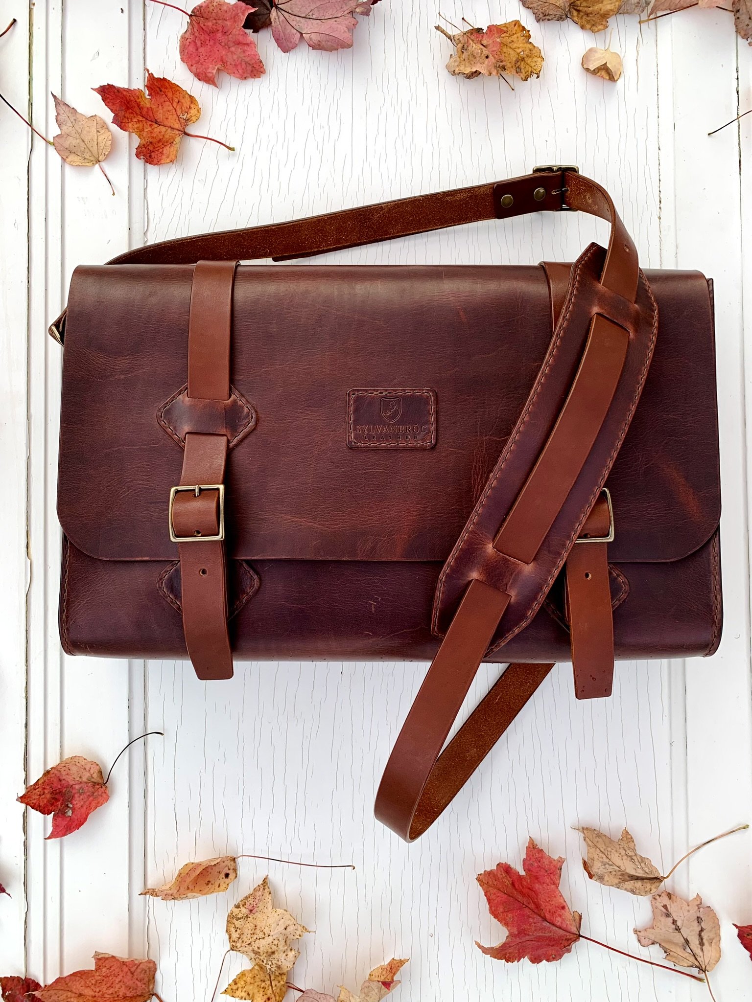 A brown leather messenger bag with straps and a buckle closure, placed on a white wooden surface with scattered autumn leaves.
