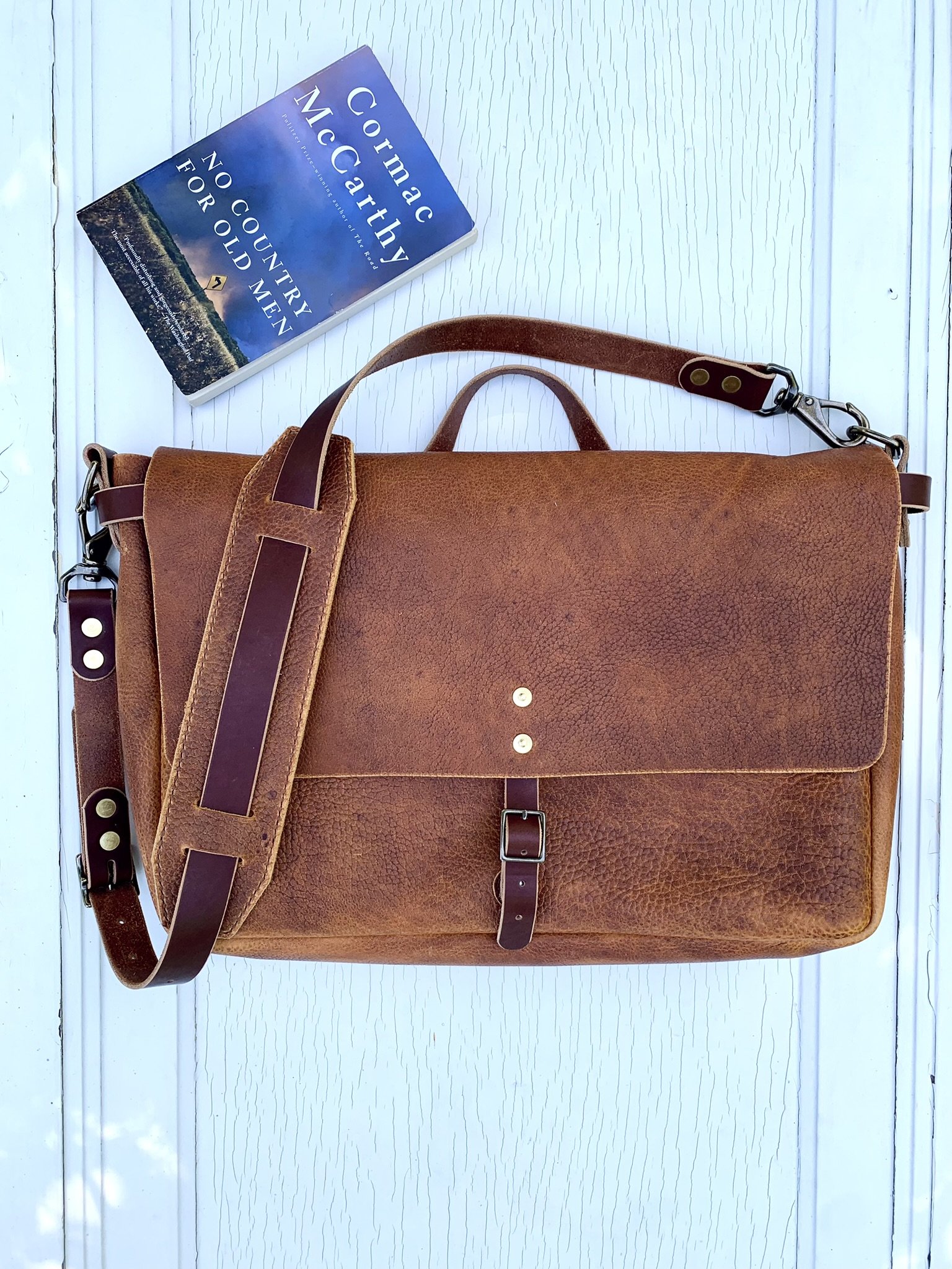 Brown leather briefcase with adjustable shoulder strap and top handle, placed on a white wooden surface; a blue book titled 'No Country for Old Men' by Cormac McCarthy is nearby.