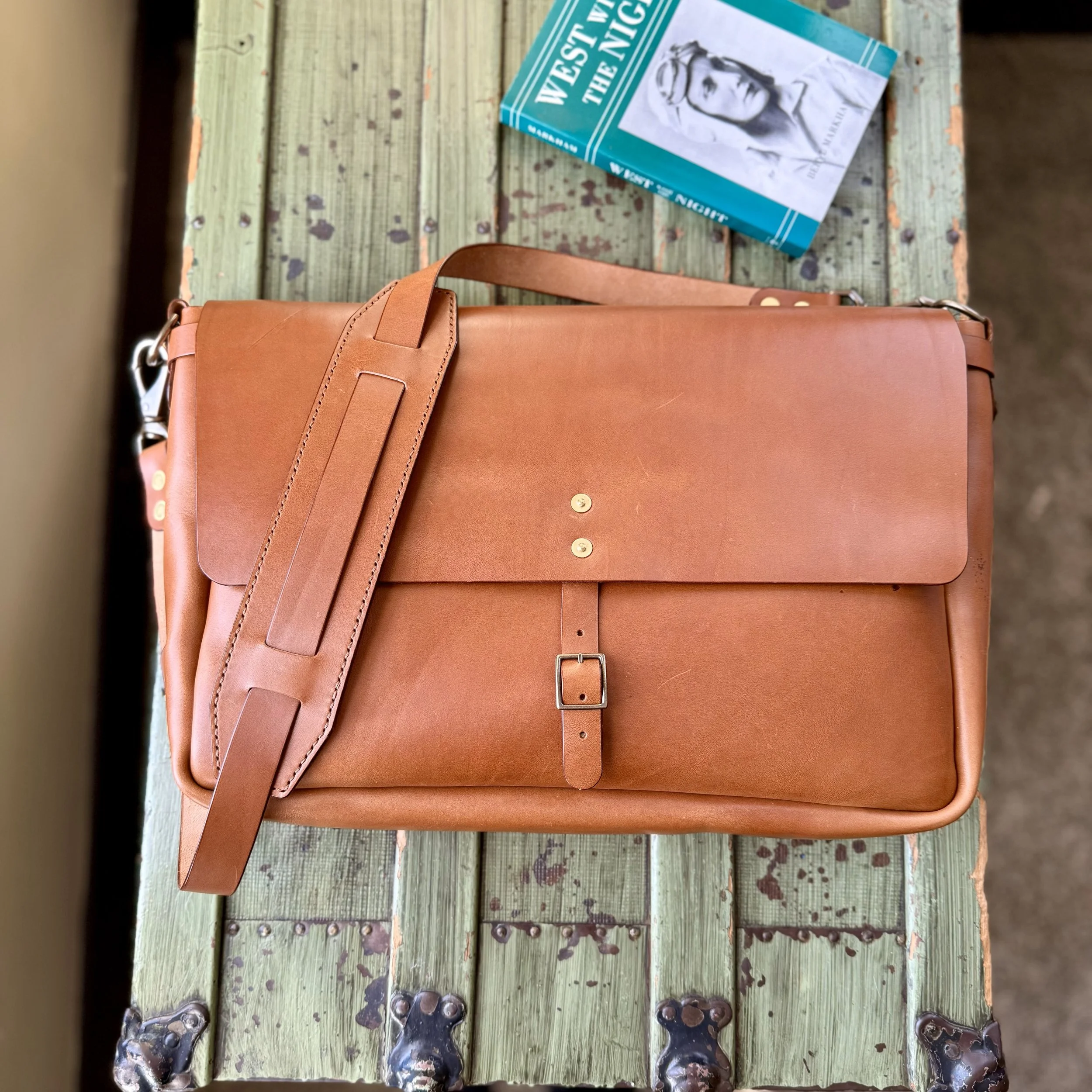 A brown leather briefcase with a strap and buckle lies on a weathered green wooden surface. A turquoise book titled 'West of the Night' is partially visible in the top right corner.