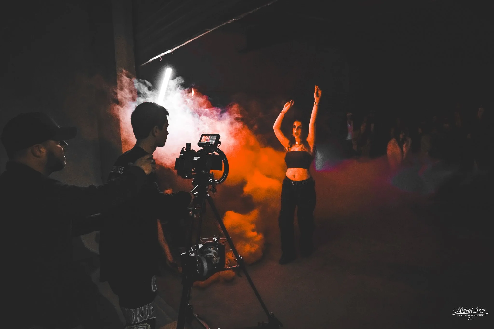 A woman dancing with her arms raised in a smoky, dark environment with colored lights, while a person with a camera films her. Other people are faintly visible in the background.