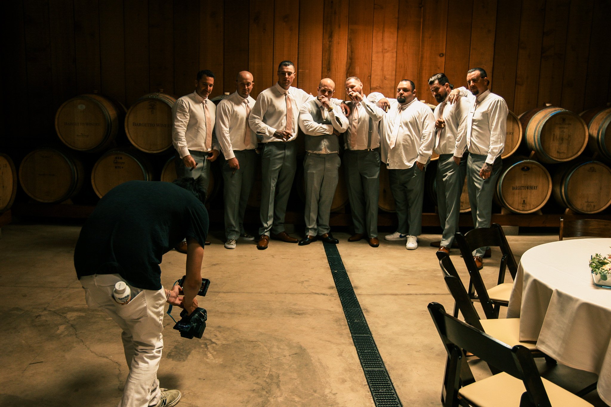 A group of nine men dressed in formal attire, standing side by side in front of wooden wine barrels at a winery, with one person in the foreground taking a photo.