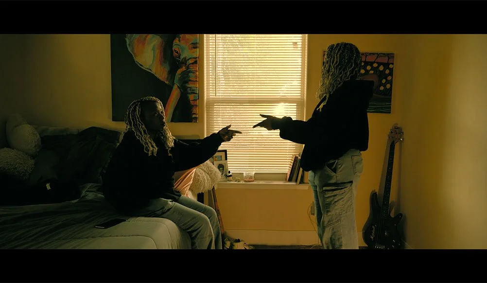 Two women with dreadlocks pointing at each other in a dimly lit bedroom, one sitting on a bed and the other standing near a window with blinds. A guitar leans against the wall, and there are framed photos and artwork in the room.